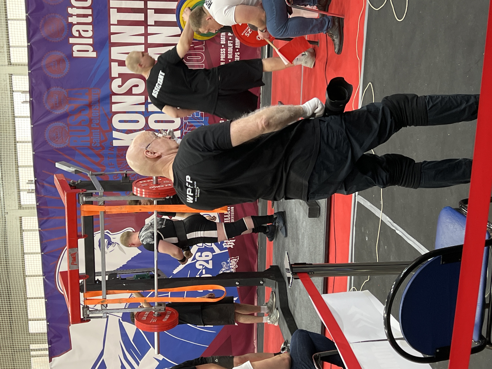 Here is a description of the image:
The image shows a powerlifting competition taking place in Saint Petersburg, Russia. The event is labeled "Konstantinov Anti". There are multiple individuals present, including competitors and assistants. One man, wearing a black shirt, has "WPFP" printed on his shirt and faces the camera. He stands to the right of the lifting rig. The lifting rig is red with orange straps and holds plates labeled “ELEIKO”. Competitors are seen lifting weights. There is a banner with the words "RUSSIA" and "Saint Petersburg" and the text "KONSTANTINI ANT" displayed in the background. A red barrier is in place to separate spectators from the competition area. There are also white and blue chairs available.