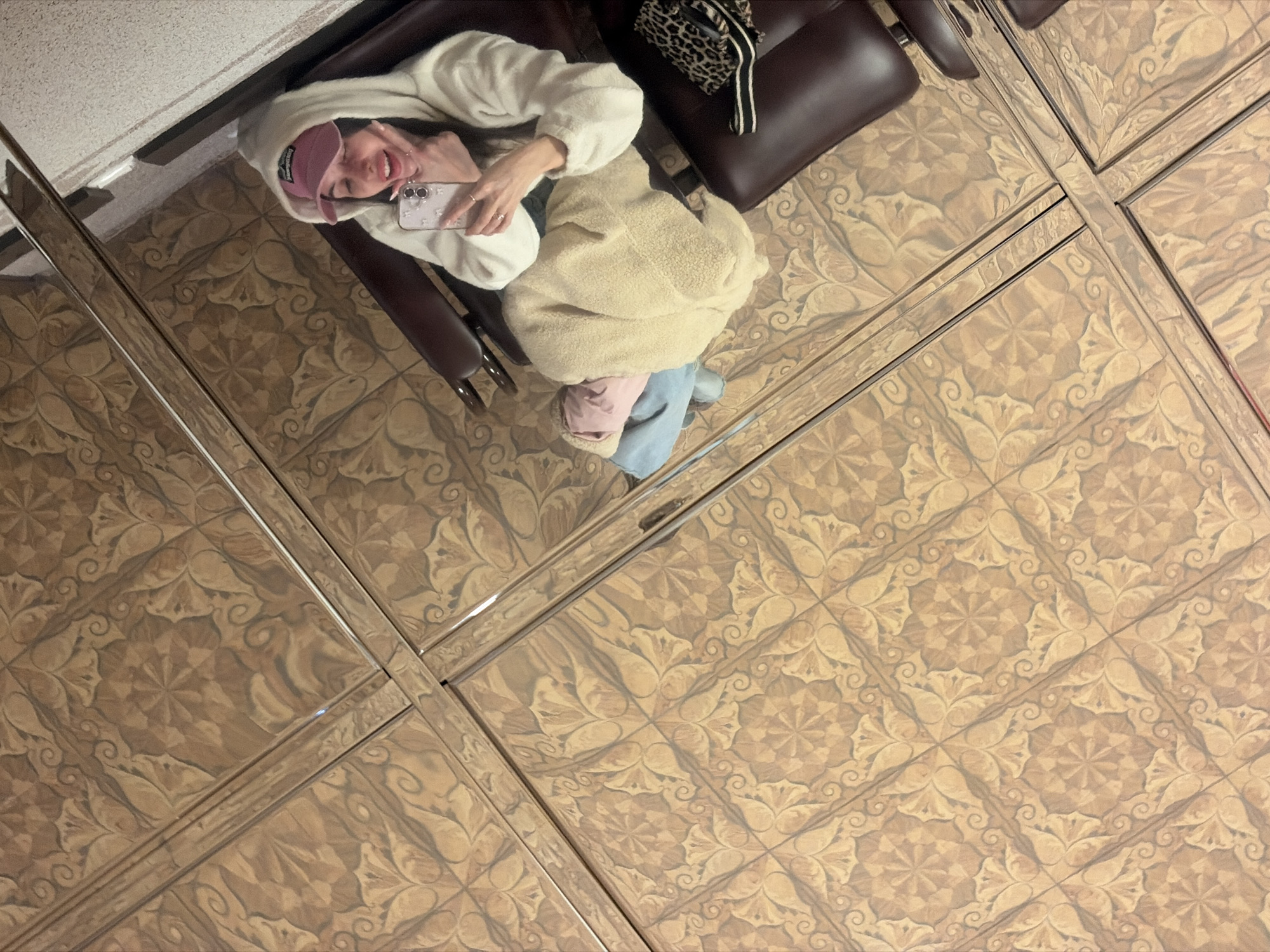 The image presents a unique mirror selfie, captured from an elevated perspective, reflecting off a highly polished and patterned floor. A person, appearing female, is seated comfortably in a dark brown leather armchair. She is dressed in casual attire, including a cream-colored hoodie, light blue jeans, and a light pink baseball cap worn backward. A soft, light beige sherpa-style jacket or blanket is draped over her lap. A leopard print purse with a striped strap rests on the chair next to her.
The person is actively taking the selfie, smiling broadly with teeth visible, and making a peace sign with her left hand. She holds a light-colored iPhone, featuring a decorative, possibly floral or starry, clear case, in her right hand. Her nails appear manicured.
The most prominent feature of the scene is the floor, which is composed of numerous square tiles displaying an intricate, repeating botanical or arabesque pattern in various shades of brown and beige. These tiles are exceptionally reflective, creating a mirror-like surface that clearly reflects the person and the overhead lighting, giving the impression of multiple reflective panels. The reflection quality of the floor is so strong that it functions as the primary mirror for the selfie. The overall mood conveyed is casual and cheerful. No visible text is present in the image. The location context provided is Saint Petersburg, Russia.
