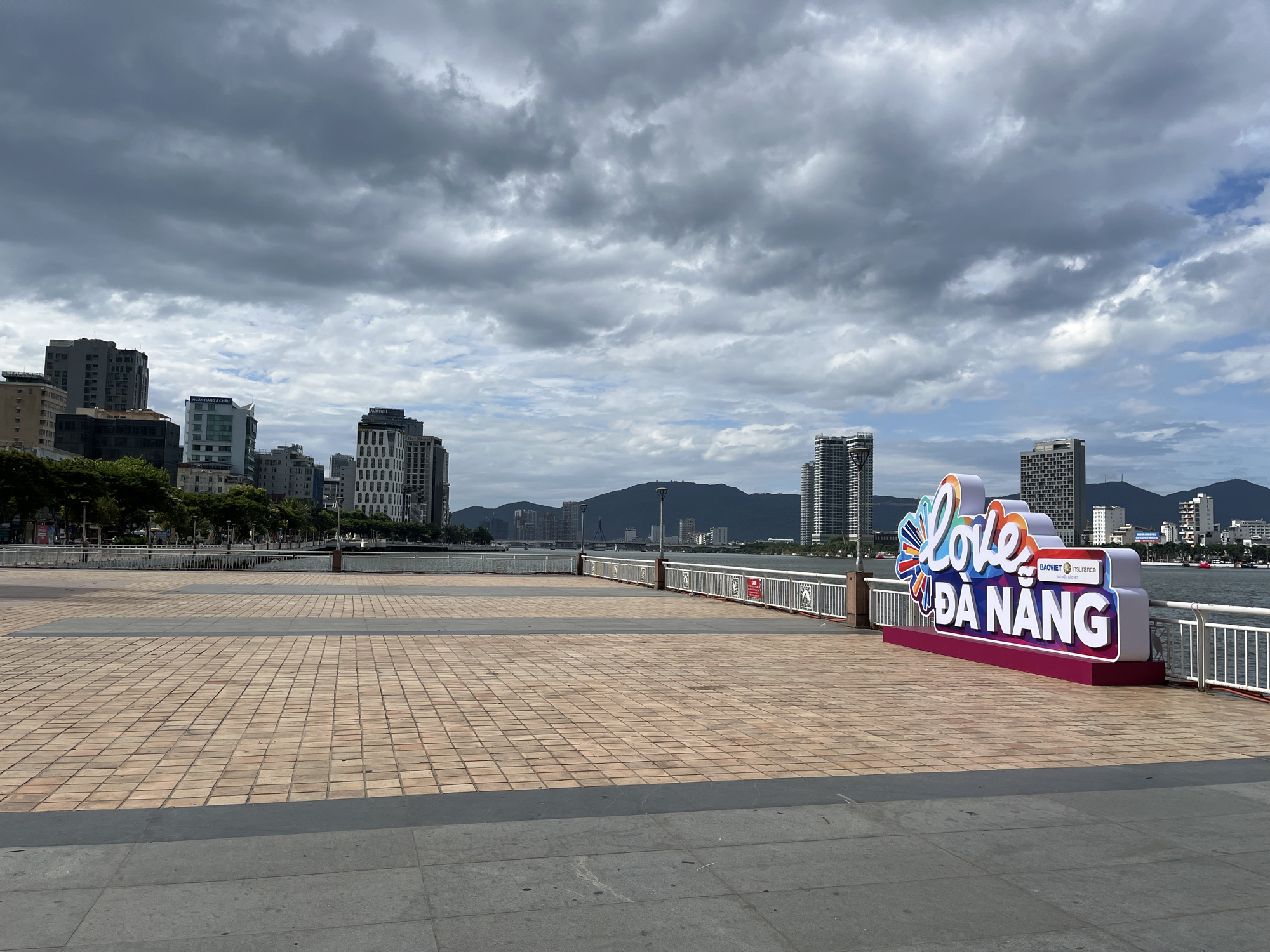 The image captures a wide, empty waterfront promenade in Đà Nẵng, Vietnam, under an overcast sky. In the foreground, a large public space paved with light brown rectangular tiles and bordered by dark grey sections stretches across the frame. A white metal railing separates the promenade from the water, which appears to be a river or bay.
Prominently featured on the right side of the image, next to the railing, is a colorful, three-dimensional sign that reads "Love ĐÀ NẴNG" in stylized, bright letters. Below the word "Love," a smaller logo for "BAOVIET Insurance" is visible.
In the mid-ground and background, the city skyline of Đà Nẵng is visible across the water. Numerous modern high-rise buildings line the shore, with some mountains providing a natural backdrop in the distance. Text visible on specific buildings includes "NGÂN HÀNG Á CHÂU" on a white high-rise and "Marriott" on another distinctive building.
The weather appears cloudy, with thick, dark grey clouds dominating the sky, though some lighter patches and hints of blue are visible towards the right. The overall light suggests it is daytime, possibly late morning or afternoon. There are no people visible on the promenade, giving the scene a calm and serene atmosphere. The presence of the "Love ĐÀ NẴNG" sign explicitly marks the location as a tourist-friendly spot in the city.