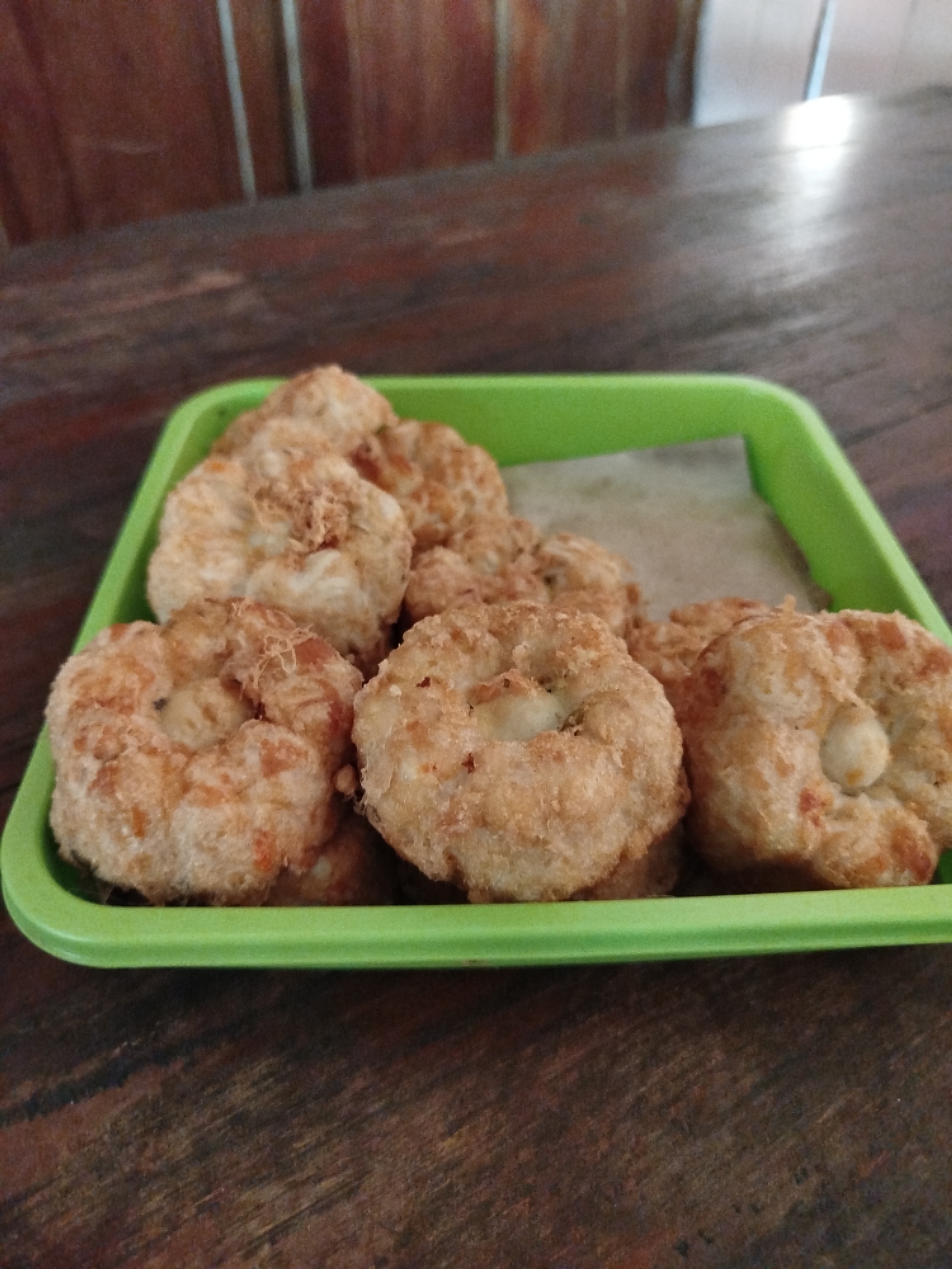 A shallow, rectangular, bright green plastic tray filled with several golden-brown, toroidal-shaped fritters sits on a dark brown wooden table. The fritters, some slightly overlapping, are textured and appear to be made of a finely ground mixture, possibly with small pieces of ingredients visible. A piece of light-colored paper is beneath the fritters within the tray. The wooden table has a visible grain pattern and appears slightly worn. In the background, out of focus, are vertical wooden slats and a bright, undefined area. The image is taken from a slightly elevated perspective, looking down into the tray. The scene is indoors, likely in a casual dining setting.