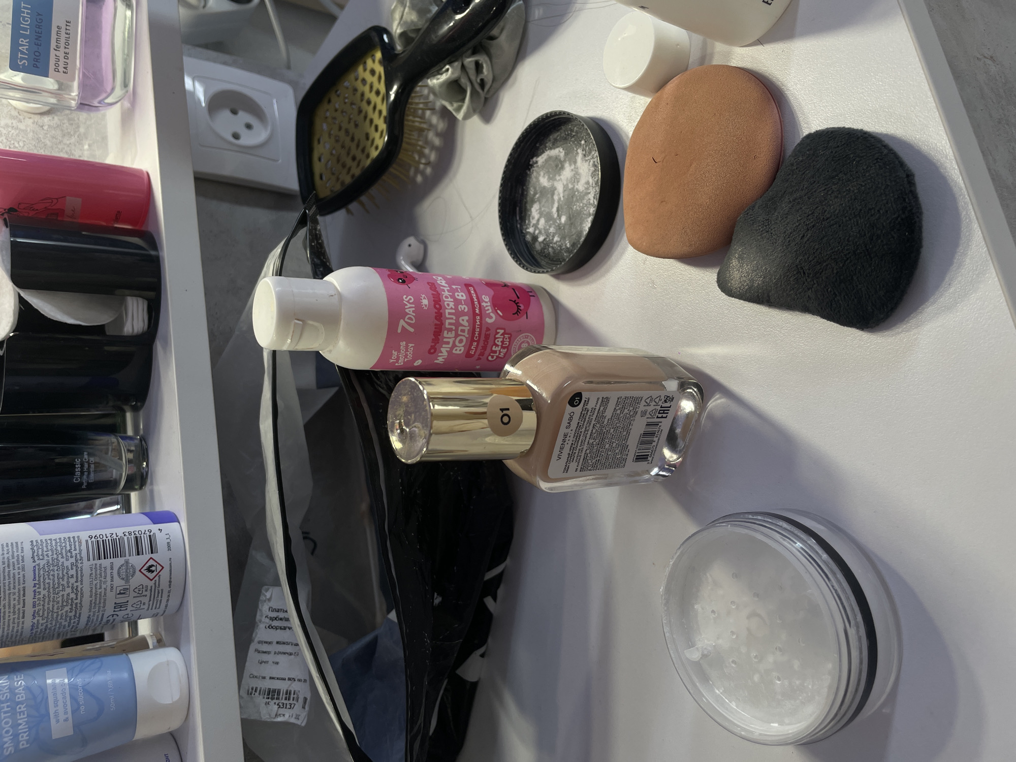 The image shows a collection of beauty products on a white surface, likely a table or shelf. The products include makeup (foundation, primer, powder), skincare (micellar water), perfume, and hair care items (hairbrush). The micellar water is labelled "7DAYS" with text in Russian. The foundation bottle is "VIVIENNE SABÓ 01". The primer is labelled "SMOOTH SKIN PRIMER BASE" with some text in Georgian. An aerosol can has Russian text, indicating it's likely a deodorant. A tag with text in Russian is attached to an unidentified dark item. The perfume bottle is labelled "STAR LIGHT PRO-ENERGY". Given the presence of products with Russian and Georgian labels, the location is likely in Russia, possibly Chaikovsky given the mention of this city in the prompt, although this cannot be confirmed from the image alone. The products appear to be arranged in a casual manner, suggesting they are in use.