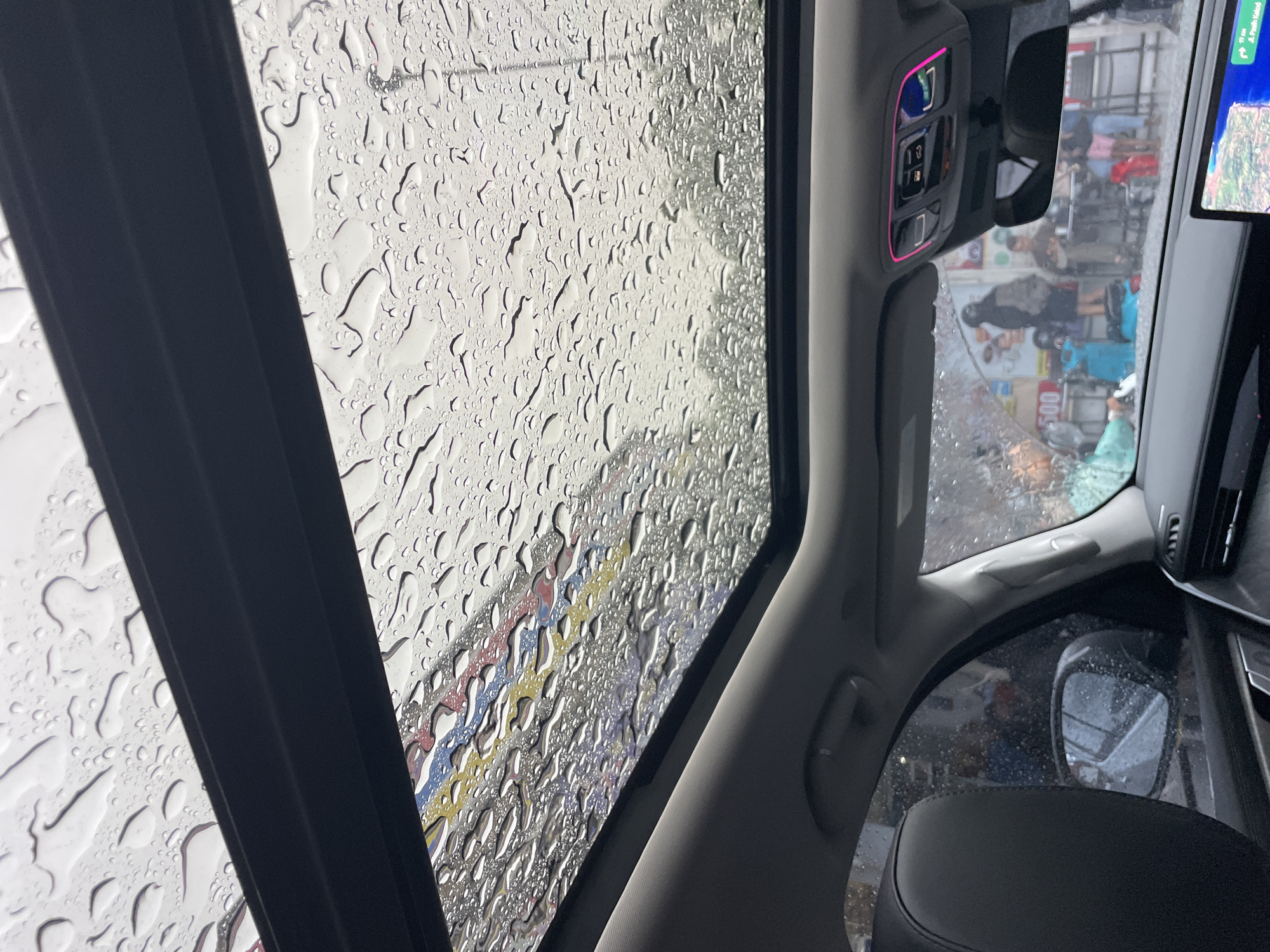 The view is from inside a car on a rainy day. The sunroof is open, and rain droplets are visible on the glass. Through the sunroof, there is a colorful banner with red, yellow, and blue stripes. The car's interior includes buttons with pink accents, a rearview mirror, a sun visor, a dashboard, and a side mirror. Outside the car, a street scene is visible with people walking and standing near buildings, possibly in a city.