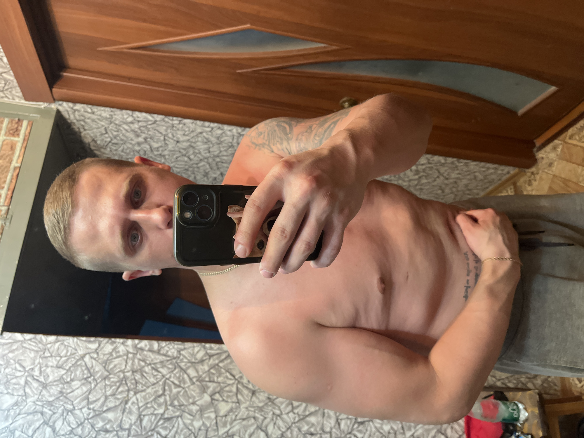 A young Caucasian male with a short haircut and light blue eyes is taking a mirror selfie indoors. He is shirtless, wearing grey sweatpants, and has a gold chain around his neck. A tattoo is visible on his left arm. His skin appears to be slightly sunburned on his shoulders and chest. He is holding a black smartphone with a case featuring a dog's image. The background shows a wall with textured wallpaper and a wooden door. A bottle of water and other items are visible on a shelf to the left. The location is identified as Minsk, Belarus.