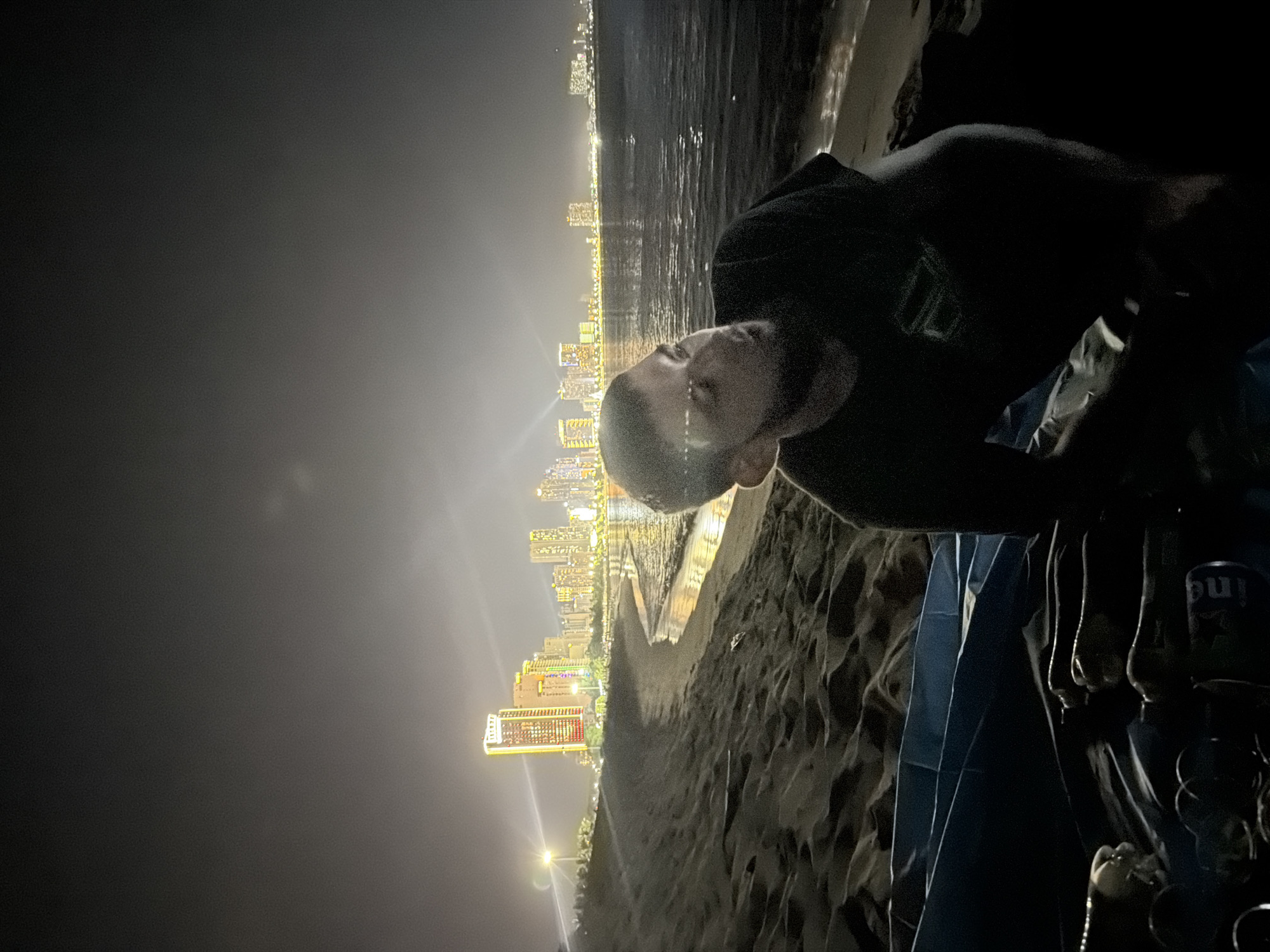A nighttime, low-angle, medium shot shows a man sitting on a beach in front of a body of water, with a city skyline illuminated in the background. The man is wearing a dark t-shirt with a logo on the left chest. He is looking to his right, away from the camera. In the foreground, partially obscured, is a blue tarp and several empty beverage cans. The beach is dark and sandy, with visible footprints. The water is calm with reflections of the city lights. The city skyline features numerous tall buildings, many of which are brightly lit. The scene is likely taking place along the coast of Nha Trang Ward, Vietnam, given the presence of a beach, city skyline, and the context provided.