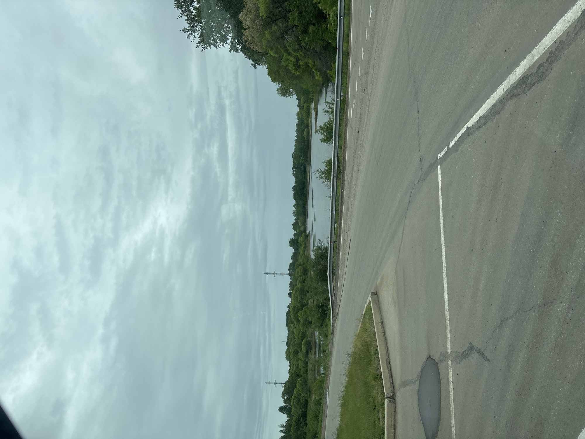 The image captures a static roadside scene alongside a body of water under an overcast sky. In the foreground, an asphalt road occupies the right side of the frame, featuring a solid white line on its right edge and a dashed white line closer to the center, along with some cracks and dark repair patches. A circular utility cover or manhole is visible on the asphalt near a concrete curb that separates the road from a strip of green grass.
A metal guardrail runs parallel to the road, separating it from a wide body of water, possibly a river or lake, that fills the midground. The water reflects the pale, cloudy sky. Dense green trees and foliage line the far bank of the water. In the distance, rising above the treeline, two distinct electrical transmission towers (pylons) are visible. The sky is uniformly covered with light grey, textured clouds, indicating an overcast day. The time of day appears to be daytime, but the cloudy conditions prevent a precise determination of morning or afternoon. There are no people, animals, or vehicles visible, and no discernible text. The overall scene conveys a calm, natural setting adjacent to infrastructure.