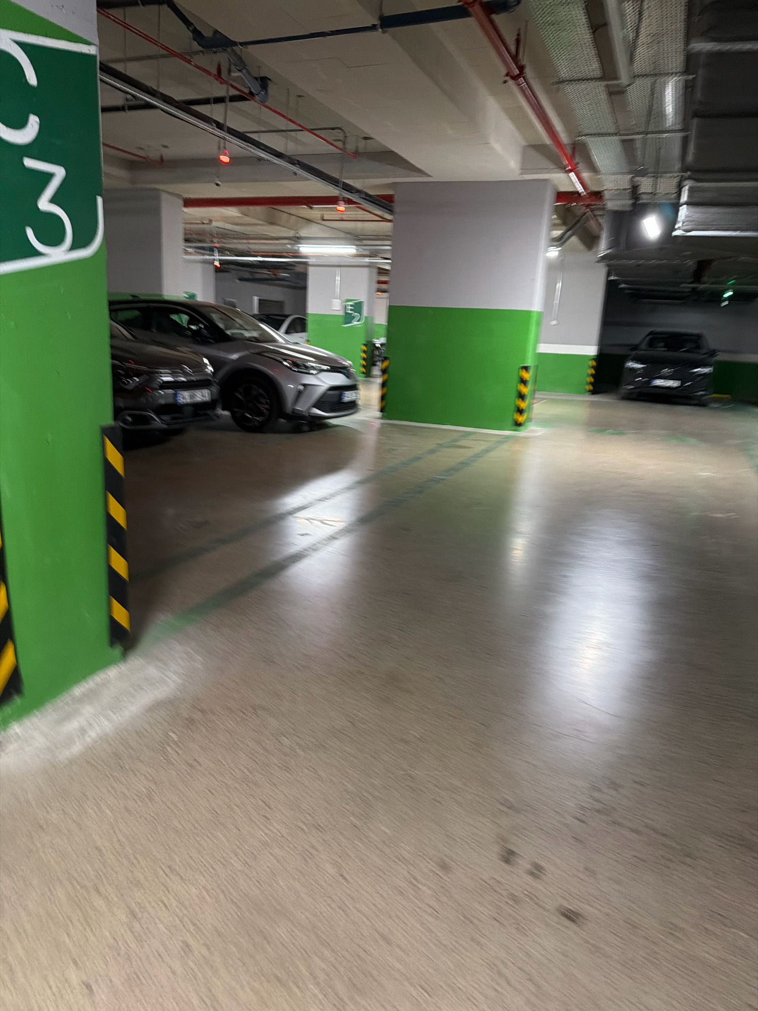 The image depicts a multi-level underground parking garage in Ataşehir, Turkey, as indicated by the green pillar with the number "3" visible on the left. The garage features concrete flooring and ceiling with exposed piping and electrical conduits. Several cars are parked in designated spots. In the foreground, a silver car and a dark-colored car are partially visible. Further back, a black SUV is parked. The walls of the garage are painted in sections of white and green, with yellow and black striped bollards placed near pillars. Lighting fixtures are visible on the ceiling. The overall impression is one of an organized, functional, and well-maintained parking facility.