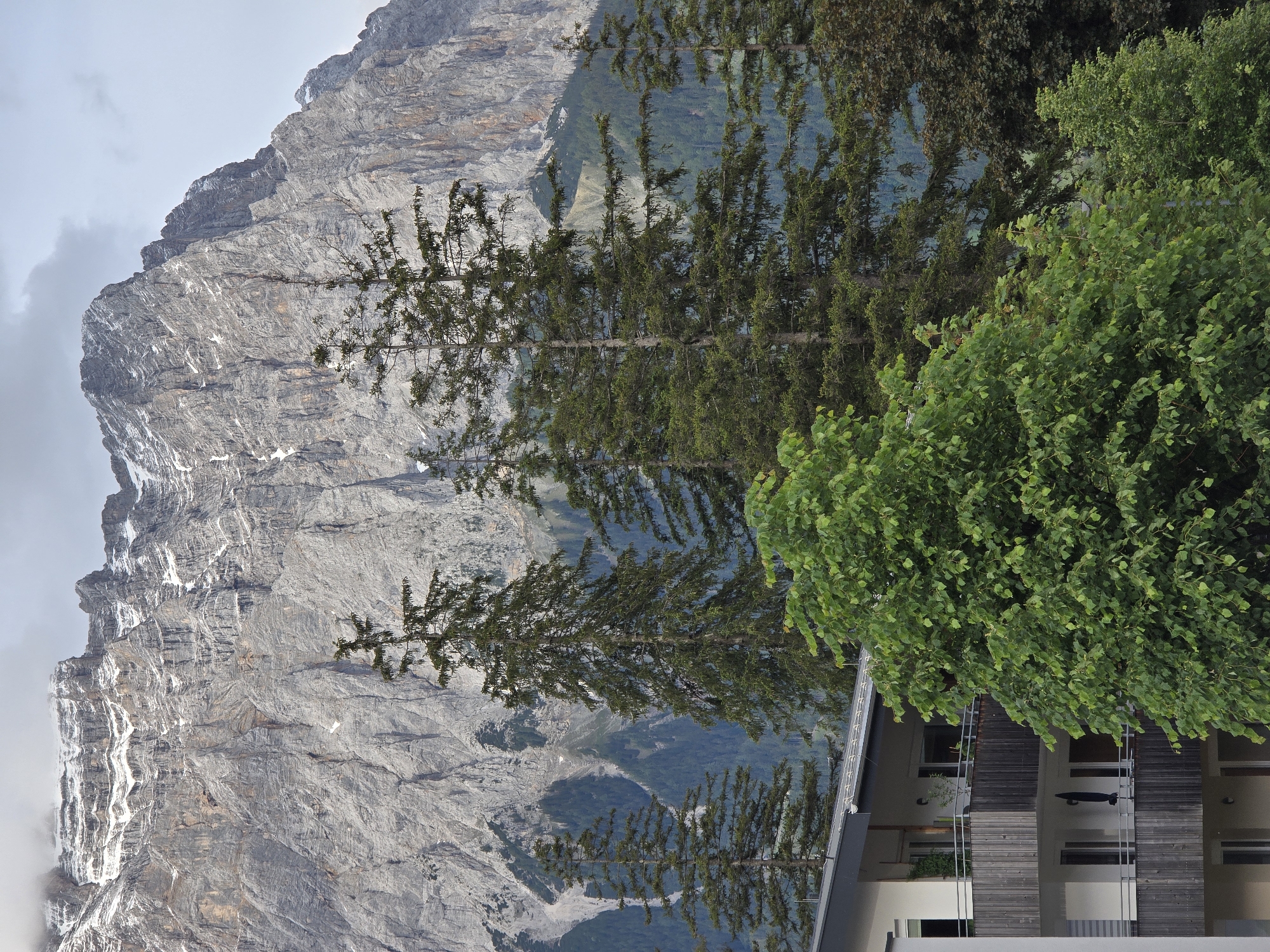 In Lermoos, Austria, a residential building with multiple balconies faces a majestic mountain range under a cloudy sky. The building, constructed with white walls and dark wooden railings on its balconies, is partially obscured by lush green trees. In the background, the imposing grey rock face of the Zugspitze mountain dominates the landscape, with patches of snow visible on its summit and slopes. Tall, dark green coniferous trees stand in front of the mountain, adding depth and texture to the scene. The overall atmosphere is serene and captures the natural beauty of the Austrian Alps.