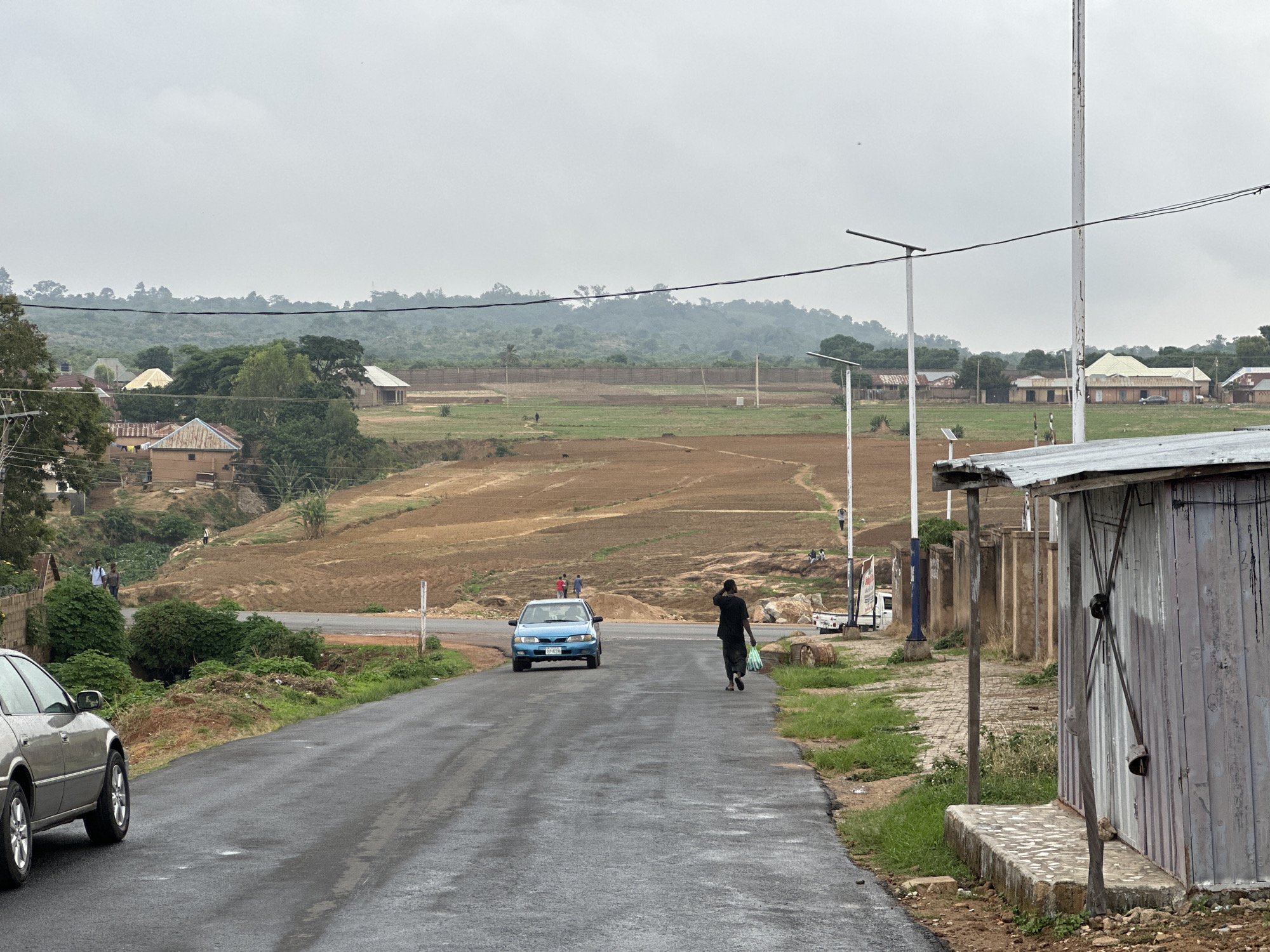 The image shows a street scene in Jos, Nigeria. The road is wet and paved, with a silver car partially visible on the left. A blue car is driving on the road towards the viewer. A person wearing black clothing walks on the right side of the road, carrying a bag. In the background, there is an open field with some trees and houses. The sky is overcast and grey. On the right, there is a weathered metal building. Utility poles stand along the roadside. People can be seen in the background near the field.