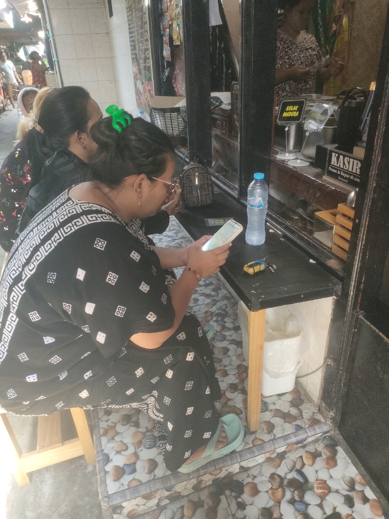 The image captures a scene outside a storefront in Surabaya, Indonesia, with two women seated at a counter. The woman in the foreground, wearing a black garment with a white geometric pattern, is looking at her smartphone. She has her hair pulled back and is wearing glasses. A green hair clip is visible in her hair. The second woman, partially visible to her left, has blonde hair and is also looking in the same direction.
Behind the counter, a sign with the text "KOLAK MADURA" is displayed. To the right, another sign reads "KASIR Pesan & Bayar di sini Terima kasih". A bottle of "Club" brand water and a set of keys with a yellow object are on the counter. In the background, other people are visible, as well as various items on display in the storefront. The flooring is a mosaic of pebbles. The lighting suggests it is daytime.