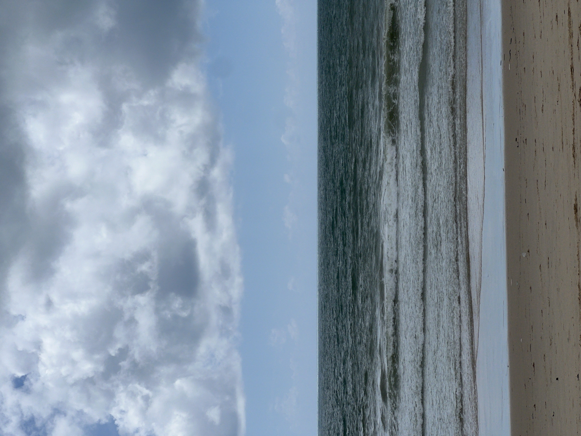 The image shows a wide shot of a beach scene with a cloudy sky and the ocean. The sky occupies the upper half of the frame, filled with large, dark gray clouds that are partially obscuring a lighter blue sky. The horizon line is sharp, dividing the sky from the ocean. The ocean is a deep, dark teal color with visible ripples and several lines of breaking waves rolling towards the shore. The foreground consists of a sandy beach. The sand is a light brown color and appears slightly wet near the water's edge, reflecting the sky. There are no people or any man-made objects visible in the image. The environment suggests a coastal setting with overcast weather. This scene could be located on a beach in Choeng Thale, Thailand, known for its coastal landscape and varying weather conditions.