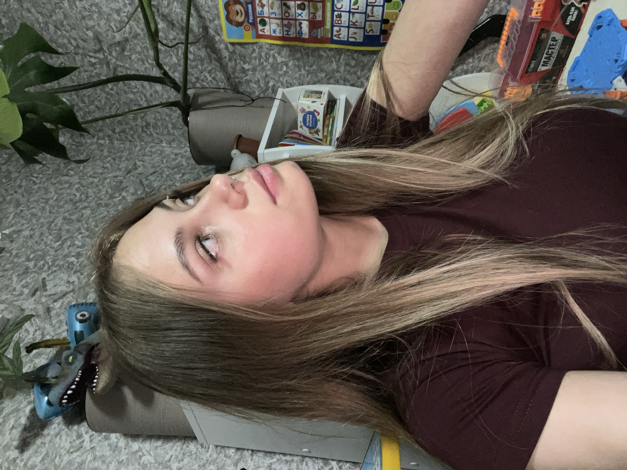 This image, captured in Фоки, Russia, depicts a young woman in an indoor setting. The woman, appearing in her late teens or early twenties, occupies the majority of the frame, positioned in the foreground. She has long, straight brown hair with blonde highlights, which falls over her shoulders. She wears a deep maroon short-sleeved t-shirt. Her head is turned slightly to her left, with her gaze directed upwards and to her right. A septum piercing is visible, and she has visible eyelashes and groomed eyebrows. Her left arm, clothed in the t-shirt, is visible in the lower left, while her right forearm is partially visible in the lower right, resting near a toy.
The background reveals a textured gray wall, likely wallpaper. Various objects are arranged on shelves and surfaces. In the upper left, a blue balloon shaped like a dinosaur head (specifically a raptor with teeth and yellow eyes) is perched on a shelf, alongside another blue inflatable object and a small green plant. To the right, a larger green-leafed plant, possibly a Monstera, sits in a gray pot on a shelf. A white shelf unit behind the woman's head holds a roll of brown material. Further to the right, another white shelf unit contains books, including one with the Russian title "ПЕРВЫЕ ЗНАНИЯ" (First Knowledge), and a white bird-shaped figurine. Dominating the far right background is a blue and yellow educational wall chart displaying the Russian alphabet ("РУССКИЙ АЛФАВИТ") with corresponding illustrations. In the lower right corner, a clear plastic toy gun with orange accents is visible, featuring Russian text "МАСТЕР" (MASTER) and "БУДЬ КАК ПАПА!" (BE LIKE DAD!). A blue and orange toy is partially visible beneath the gun.