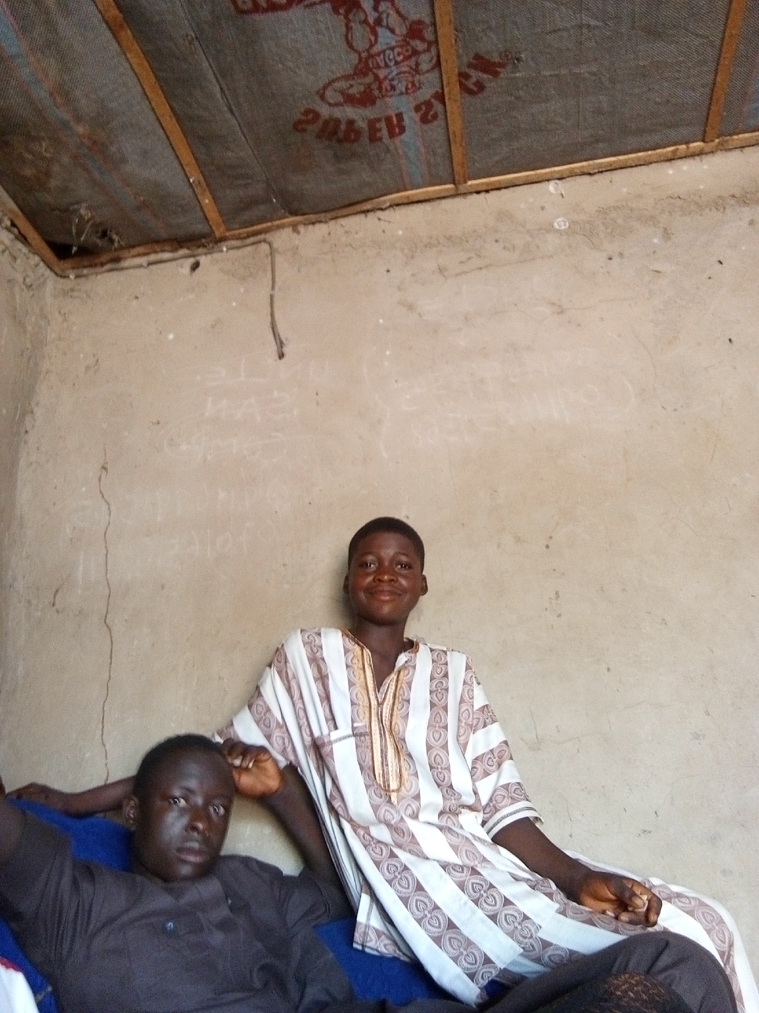 The image depicts two young Nigerian men indoors in Nyabe, Nigeria. The man in the foreground, wearing a dark brown tunic, is seated with his head tilted back and his right arm resting behind him. The second man, standing to his right, is wearing a white and brown striped garment. Both appear to be of African descent. The environment is a simple, sparsely furnished room with concrete walls and a corrugated metal roof supported by wooden beams. There are faint chalk markings or writings on the wall. The lighting is natural, suggesting daylight filtering into the space.