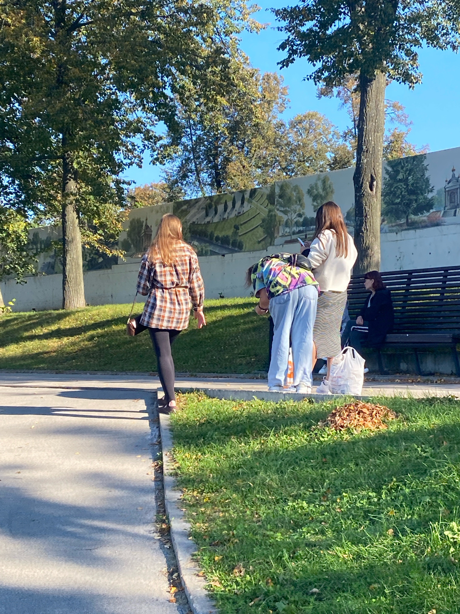 The image depicts an outdoor scene on a sunny day. Three individuals are visible. A woman with long, reddish-blonde hair, wearing a plaid dress and black leggings, walks away from the camera. To her left, another woman with long brown hair, wearing a white sweatshirt and a striped skirt, stands facing away from the camera, looking down at her phone. Beside her, a third person, dressed in a colorful, patterned shirt and light blue pants, bends over. A woman with dark hair, wearing a dark jacket and white pants, sits on a park bench in the background, appearing to observe the others. The environment includes green grass, trees with autumn foliage, a paved walkway, and a mural on a wall. A pile of fallen leaves is visible on the grass. The image does not provide any specific information about the city of Perm, Russia.