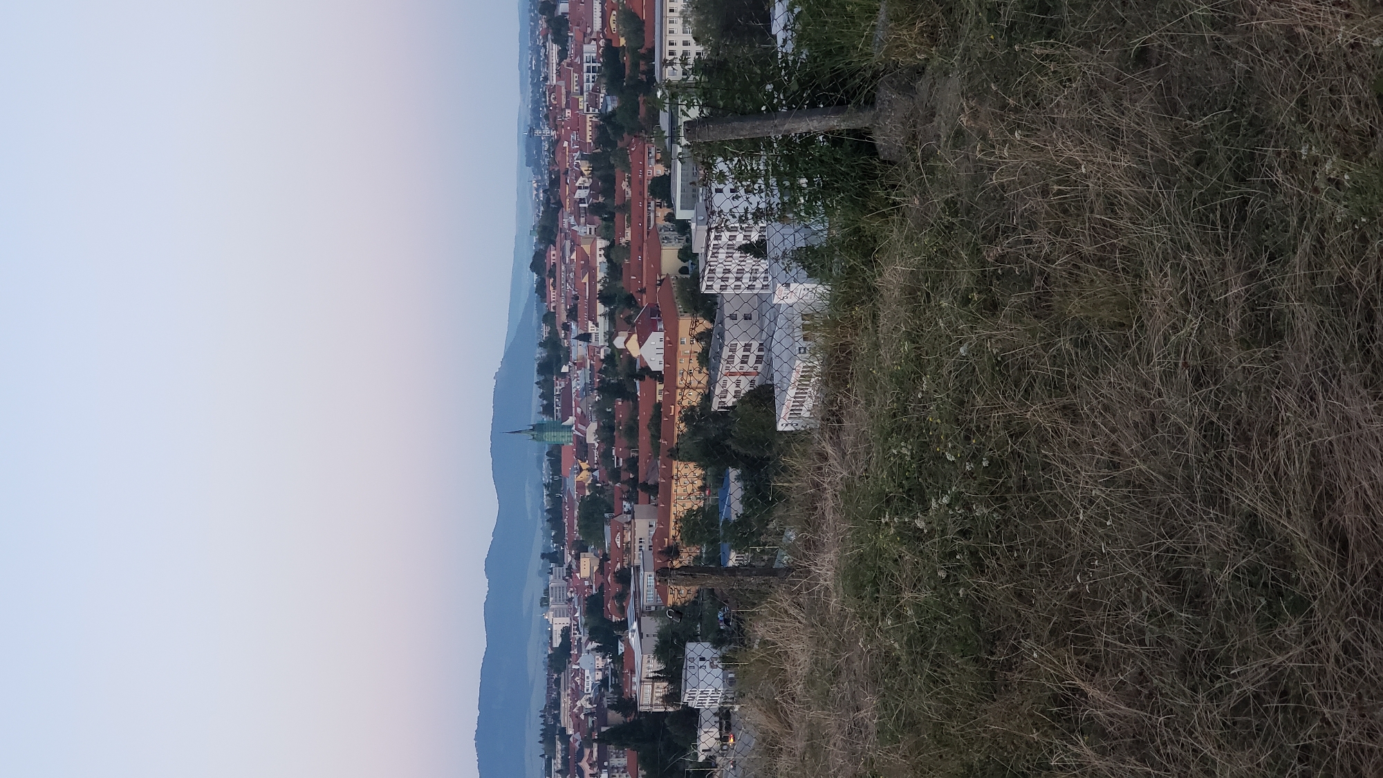The image depicts a panoramic view of the city of Prešov, Slovakia, from a slightly elevated position overlooking the urban landscape. In the foreground, dry grass and weeds grow in abundance, partially obscuring a chain-link fence supported by wooden posts. Beyond the fence, the city unfolds with a dense collection of buildings, many featuring red-tiled roofs. A prominent green spire, characteristic of a church or cathedral, rises above the cityscape, serving as a focal point. The background is dominated by rolling hills or mountains, softened by the hazy light of either dawn or dusk. The sky transitions from a pale blue at the top to a soft pink or lavender hue closer to the horizon. The overall scene conveys a sense of calm and quiet, with no people or discernible activity visible. The image appears to be a general landscape shot capturing the architectural characteristics of Prešov against a natural backdrop.