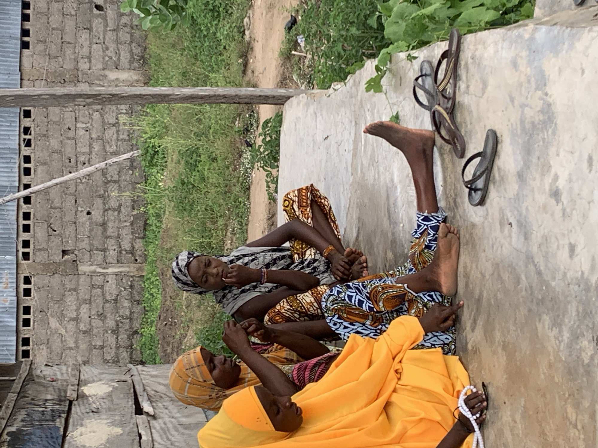 Three women are seated on a concrete surface. One woman is wearing an orange headscarf and garment, holding a string of white beads. Another woman is wearing a patterned headscarf and garment. The third woman's clothing includes blue and orange patterns. Two pairs of sandals are resting on the concrete near their feet. A cinder block wall and greenery are in the background. The location is possibly Adankolo, Nigeria.