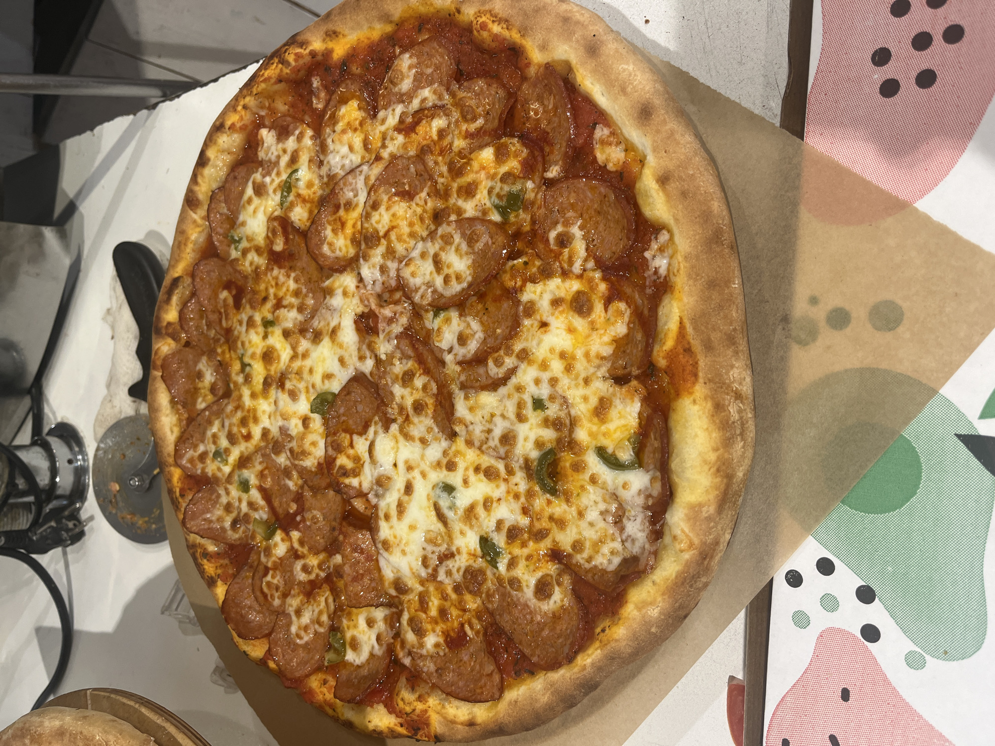A whole, round pizza topped with melted cheese, pepperoni, and what appear to be green pepper slices sits on a piece of brown paper. The pizza has a golden-brown crust that is slightly puffed and browned in places. The cheese is melted and bubbly, with some areas browned from the oven. The pepperoni slices are layered on top of the cheese and sauce, and some are curled from the heat. Small green pieces, likely from peppers, are scattered across the pizza.
The pizza is placed on a light-colored surface, possibly a table, and a piece of patterned paper with abstract shapes and dots in pink, green, and black is partially visible to the right of the pizza. To the left of the pizza, a pizza cutter with a metal wheel and black handle is visible, along with some black cords that might belong to kitchen equipment. Another round, golden-brown food item, possibly another pizza or bread, is partially visible in the bottom left corner, in a wooden basket.
The image appears to be taken indoors, in what could be a kitchen or dining area. The lighting suggests it could be daytime or evening. There is no visible text in the image. The overall impression is of a freshly prepared and appealing pizza, ready to be served or eaten. The setting seems casual, possibly in a restaurant or home.