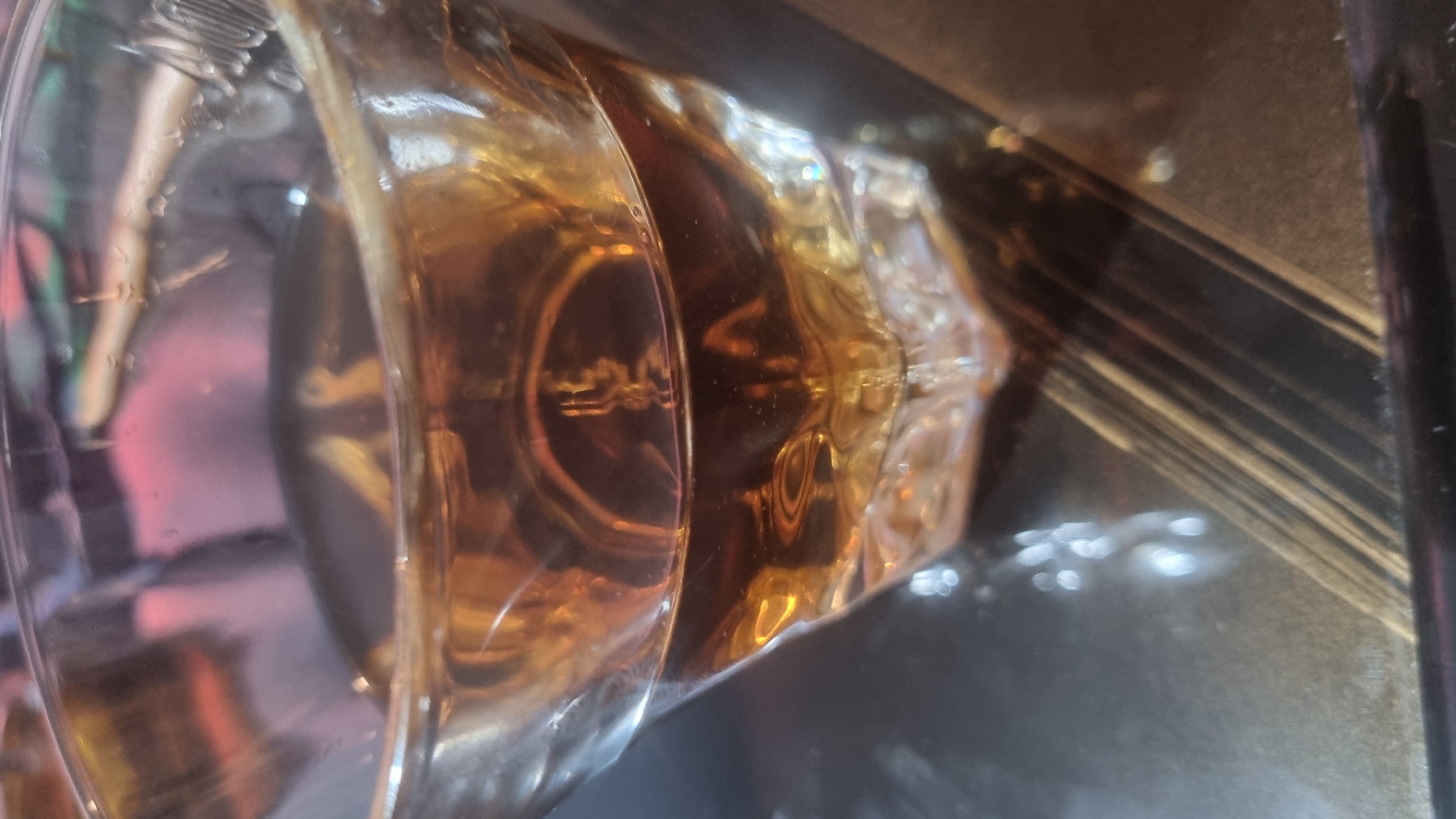 The image shows a close-up view of a glass containing a light brown liquid, likely whiskey or bourbon, sitting on a dark, reflective surface with linear patterns. The glass itself has a thick base and a rounded body. The liquid inside creates optical distortions, affecting the view of objects behind the glass. The environment appears to be indoors, but the exact location and any activities occurring are unknown based on the image.