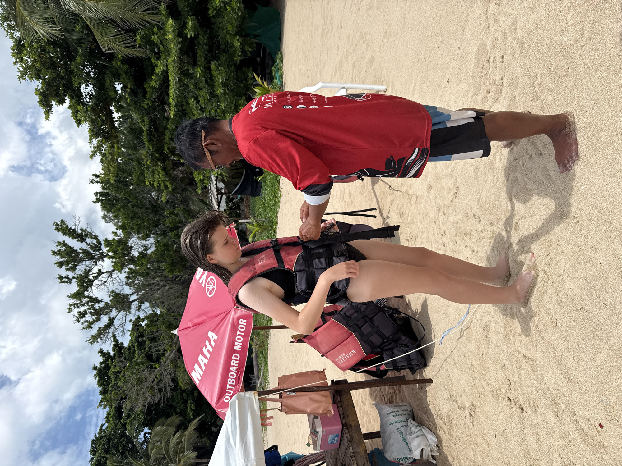 This media file depicts two people on a sandy beach, likely in Ko Samui, Thailand, given the visible text. The primary subject is a young woman in a black swimsuit and a red and black life vest, sitting on the sand. A man, wearing a red t-shirt with black and white accents and patterned shorts, is standing and appears to be adjusting or securing her life vest. A pink umbrella with "MAHA OUTBOARD MOTOR" printed on it is set up behind the woman, providing shade. Next to her is a backpack with a red and black life vest on top, and a cooler. Other items like bags and a white sheet are also present on the sand. The scene suggests preparation for a water activity, possibly jet skiing or boating, given the life vests and the "Outboard Motor" text. The background shows lush green trees and a cloudy sky, indicating it's daytime. The overall mood seems relaxed and focused on the activity.