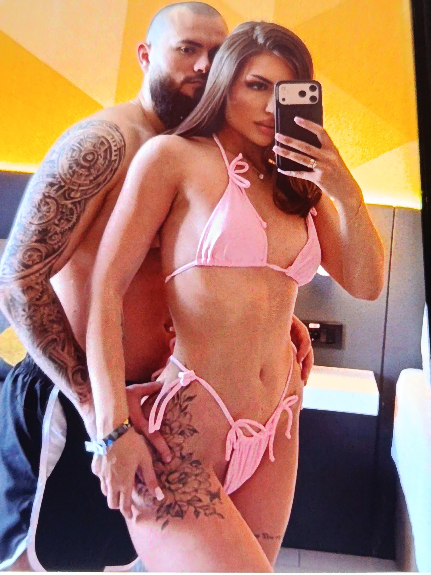 A couple is taking a selfie in what appears to be a hotel room with yellow walls. The man, to the left, has a shaved head, a beard, and is shirtless, displaying a large tattoo on his left arm. He is standing behind the woman. The woman, in the foreground, is wearing a pink bikini and holding a phone to take the picture. She has long brown hair, is wearing makeup, and has a tattoo on her thigh. Her left hand is visible, wearing a ring and holding the phone. The background shows a wall with a yellow pattern and what looks like a bed or couch. There is also a light switch on the wall. The couple appears to be embracing or posing closely for the photo. The image is taken from a slightly low angle. The location in Bălți, Moldova is not determinable from the image content.