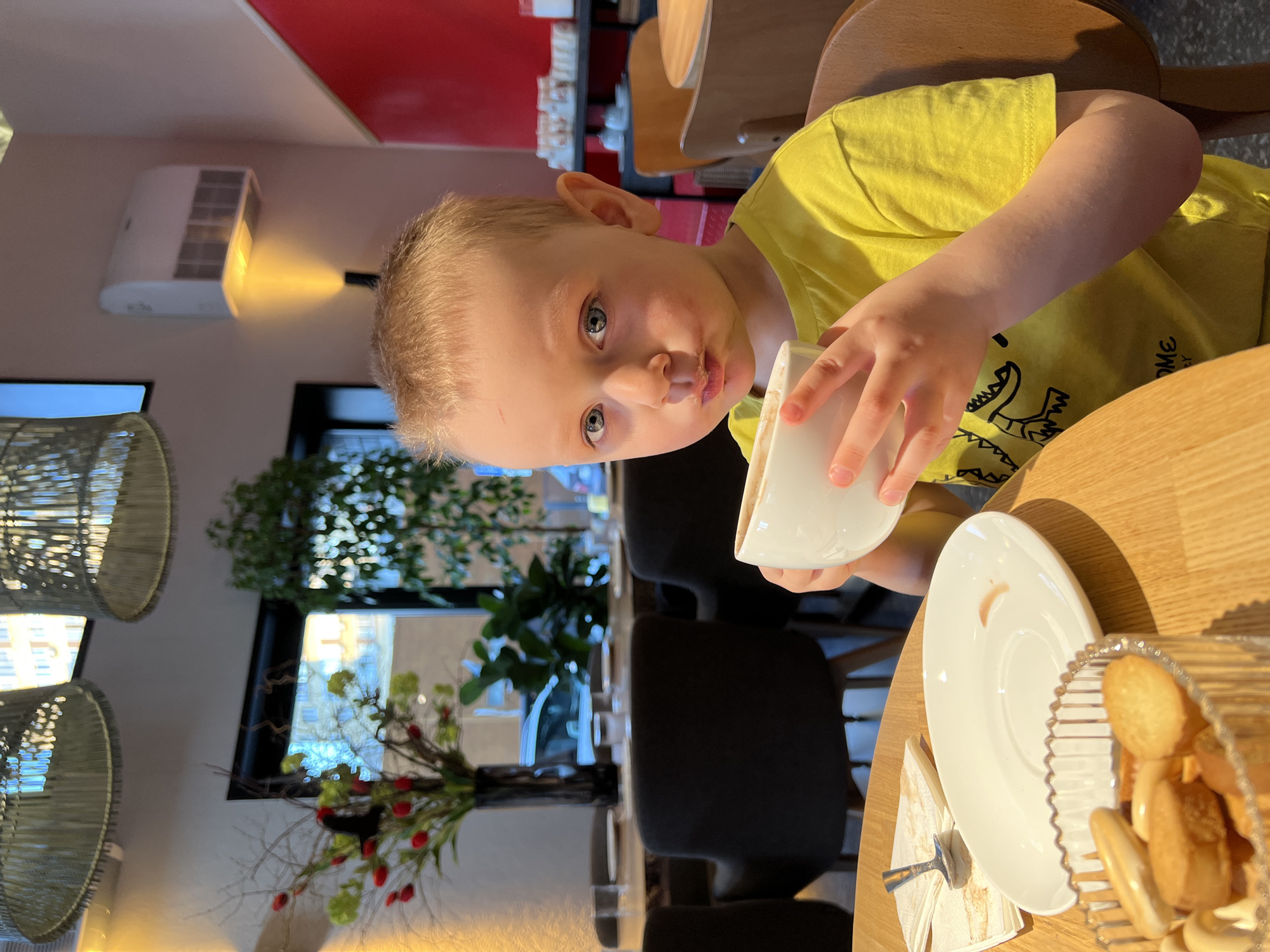 A child is sitting at a table in what appears to be a cafe. The child is wearing a yellow t-shirt and holding a white mug. On the table, there is a plate, a napkin with a spoon, and a basket of breadsticks. In the background, there are chairs, tables, a plant, and a window. The cafe is illuminated with warm light. The location is Mytishchi, Russia.