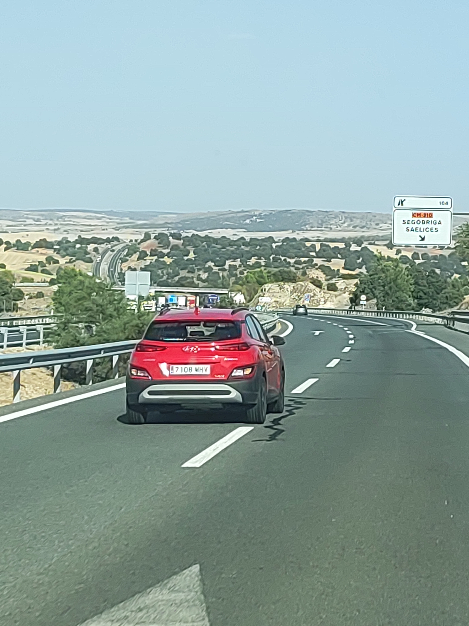The image depicts a view from a vehicle traveling on a highway in a rural landscape. The foreground is dominated by the rear of a red Hyundai Kona. The car has a Spanish license plate that reads "B7108 MHV." The highway curves to the right and extends into the distance, with other vehicles visible ahead. To the left of the highway, there is a metal guardrail and a densely vegetated hillside. In the background, a vast expanse of rolling hills with scattered trees and fields stretches towards a hazy horizon under a clear blue sky. A road sign with the text "CM-310 SEGOBRIGA SAELICES" and an arrow pointing downwards is visible on the right side of the highway, indicating directions to Segobriga and Saelices, Spain. The overall scene suggests a sunny day, likely in the summer, given the dry, arid appearance of the landscape.