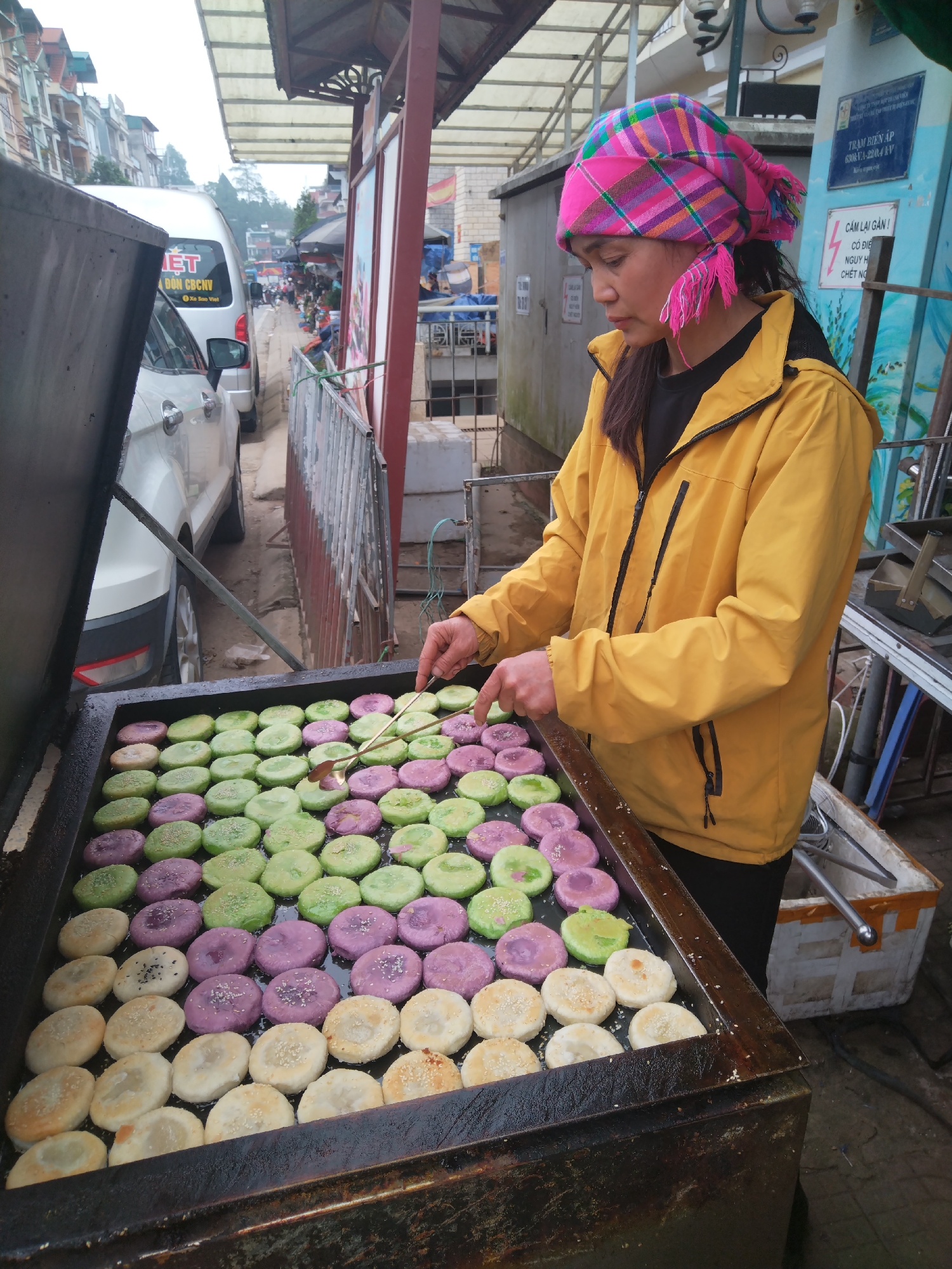 A vendor cooks colorful cakes on a flat-top grill outdoors in a city in Vietnam. A woman with dark hair wearing a bright pink plaid head wrap with tassels and a yellow jacket cooks rows of circular cakes on a large flat-top grill. The cakes are purple, green, and white, and they have sesame seeds on top. She is holding a metal utensil and appears to be moving the cakes around on the grill. Behind the grill is a sidewalk, a road, and a row of buildings. A white van is parked next to the sidewalk. The scene appears to be a busy street market. In the background there is signage in Vietnamese.