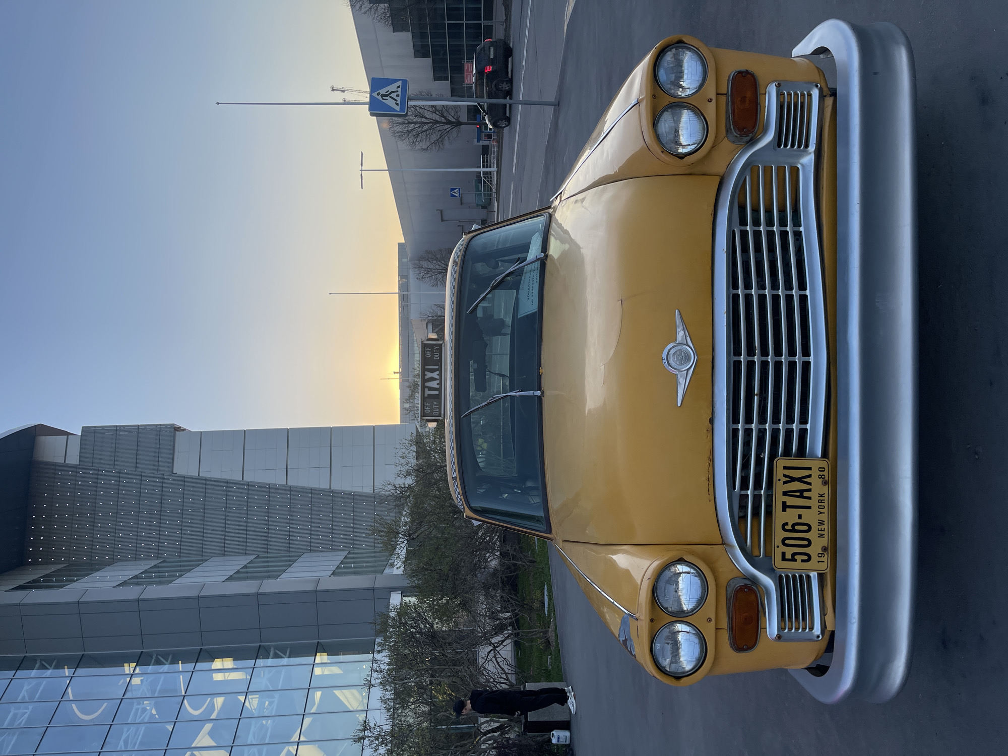 The image shows a yellow vintage Checker taxi with a New York license plate "506-TAXI" from 1980 in Moscow, Russia. The taxi is parked on a paved area in front of a modern building with glass and metal facade. A person is standing near some bushes behind the taxi. In the background, there is a road with a pedestrian crossing sign, a streetlight, and a dark car. The sky is visible in the background, appearing bright, likely during sunrise or sunset.
