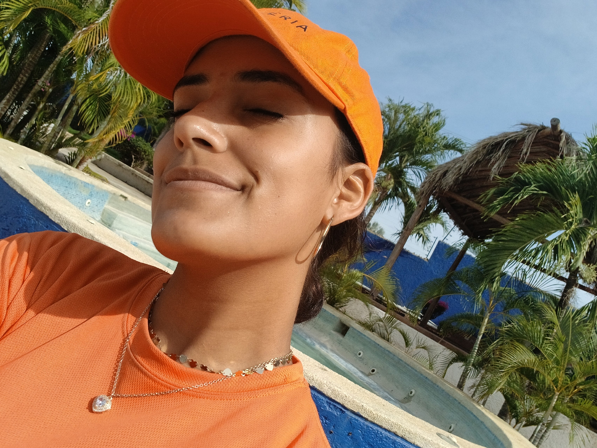 The image shows a woman wearing an orange t-shirt and cap. The woman is positioned in front of what seems to be a pool at a tropical resort. There are palm trees in the background as well as part of a thatched-roof structure. The location is consistent with Playa El Agua, Venezuela, as it features tropical vegetation and pool or resort-like structures. The woman has her eyes closed and a slight smile on her face. She is wearing a necklace with a heart-shaped pendant and gold hoop earrings.