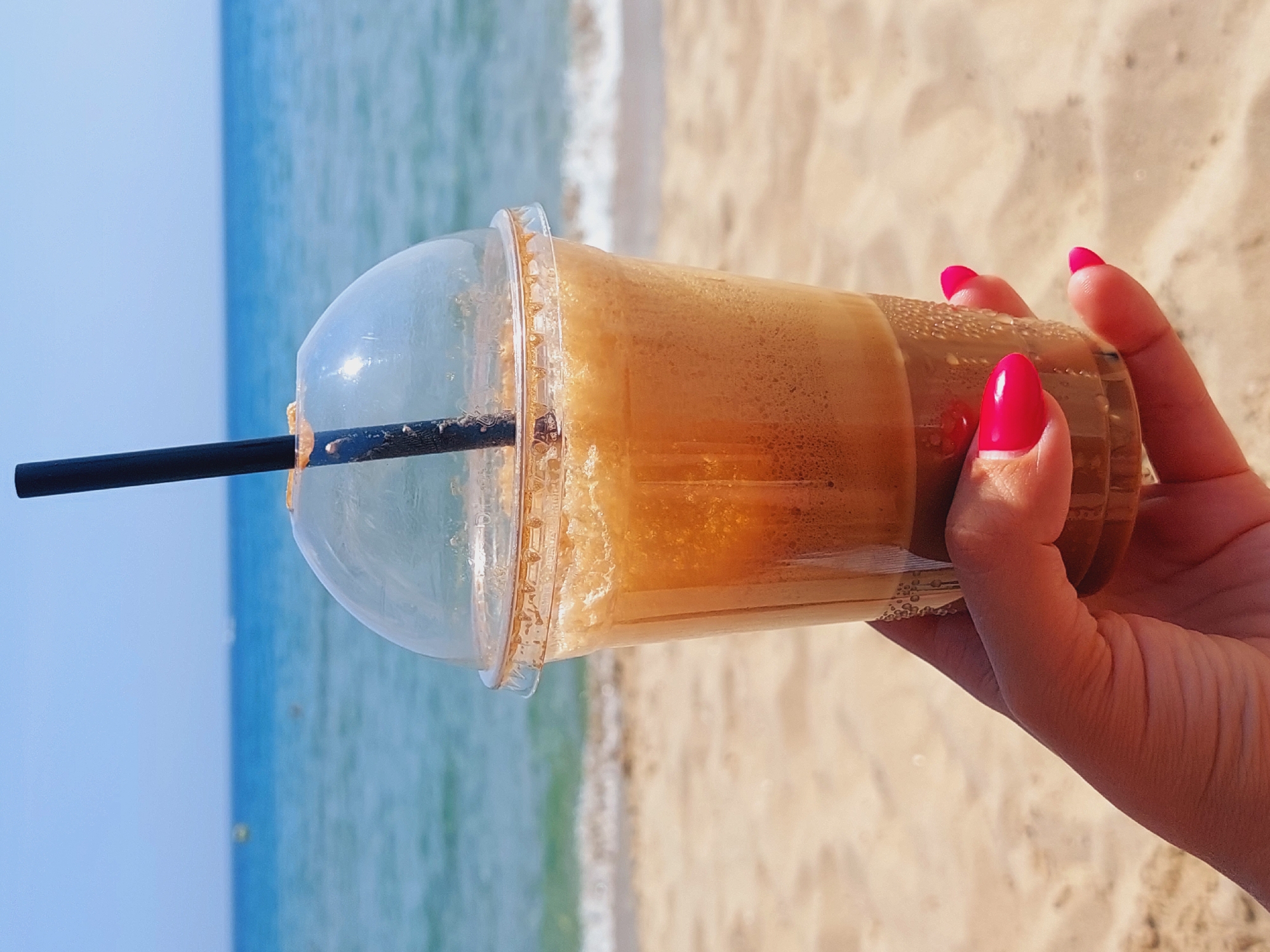 A hand with bright pink fingernails holds a clear plastic cup filled with a frothy, iced coffee beverage. The cup has a domed lid and a black straw. The background shows a sandy beach with gentle waves lapping at the shore under a clear blue sky. The overall scene suggests a relaxed vacation setting. This image is likely from the city of Sozopol, Bulgaria, known for its beautiful beaches along the Black Sea coast.