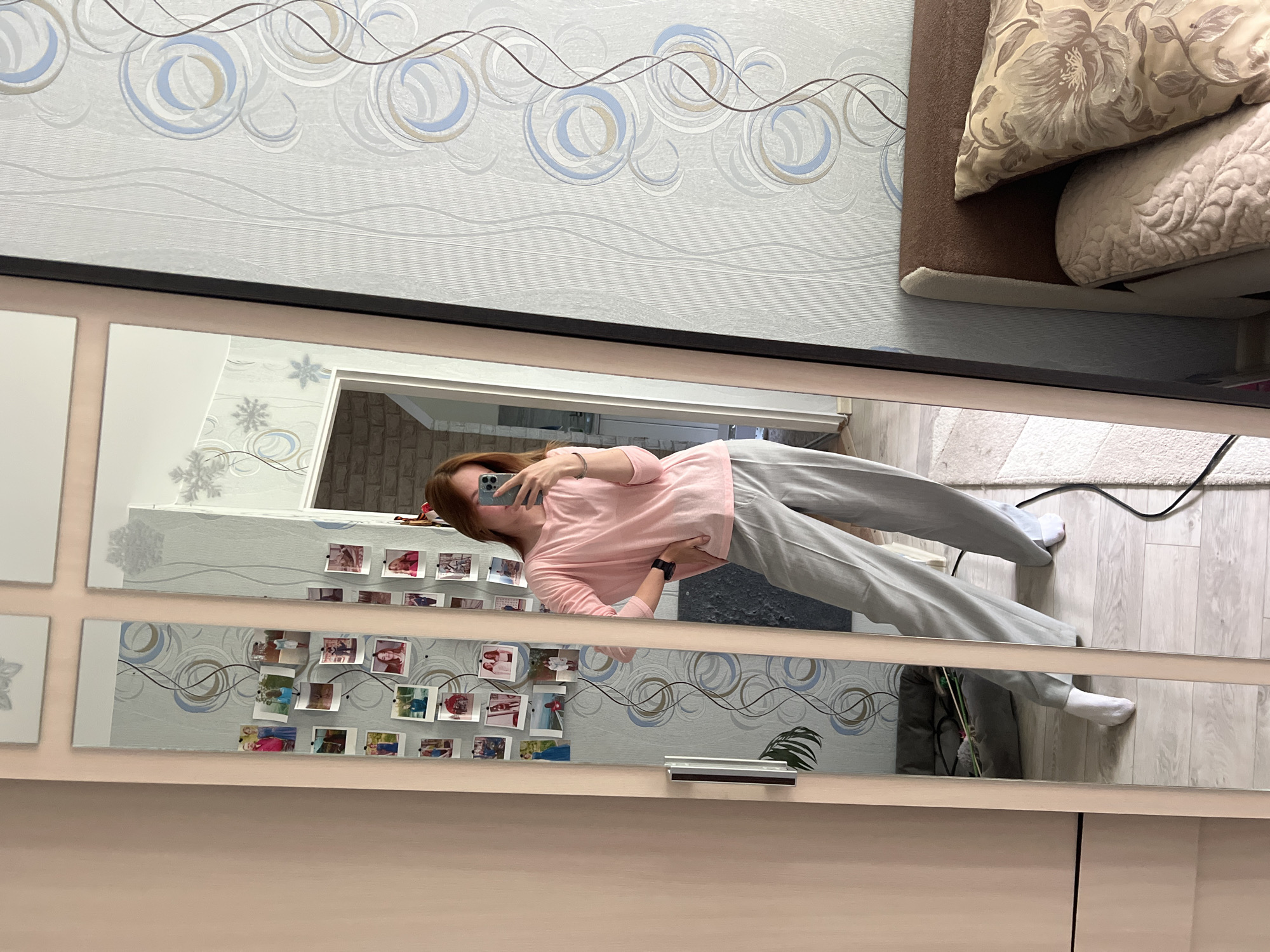 A woman in a pink top and gray sweatpants is taking a mirror selfie indoors. The mirror reflects a room with patterned wallpaper, photographs attached to the wall, and a glimpse of a doorway. The floor is light-colored wood. The location is Chaikovsky, Russia, as indicated by other available information.