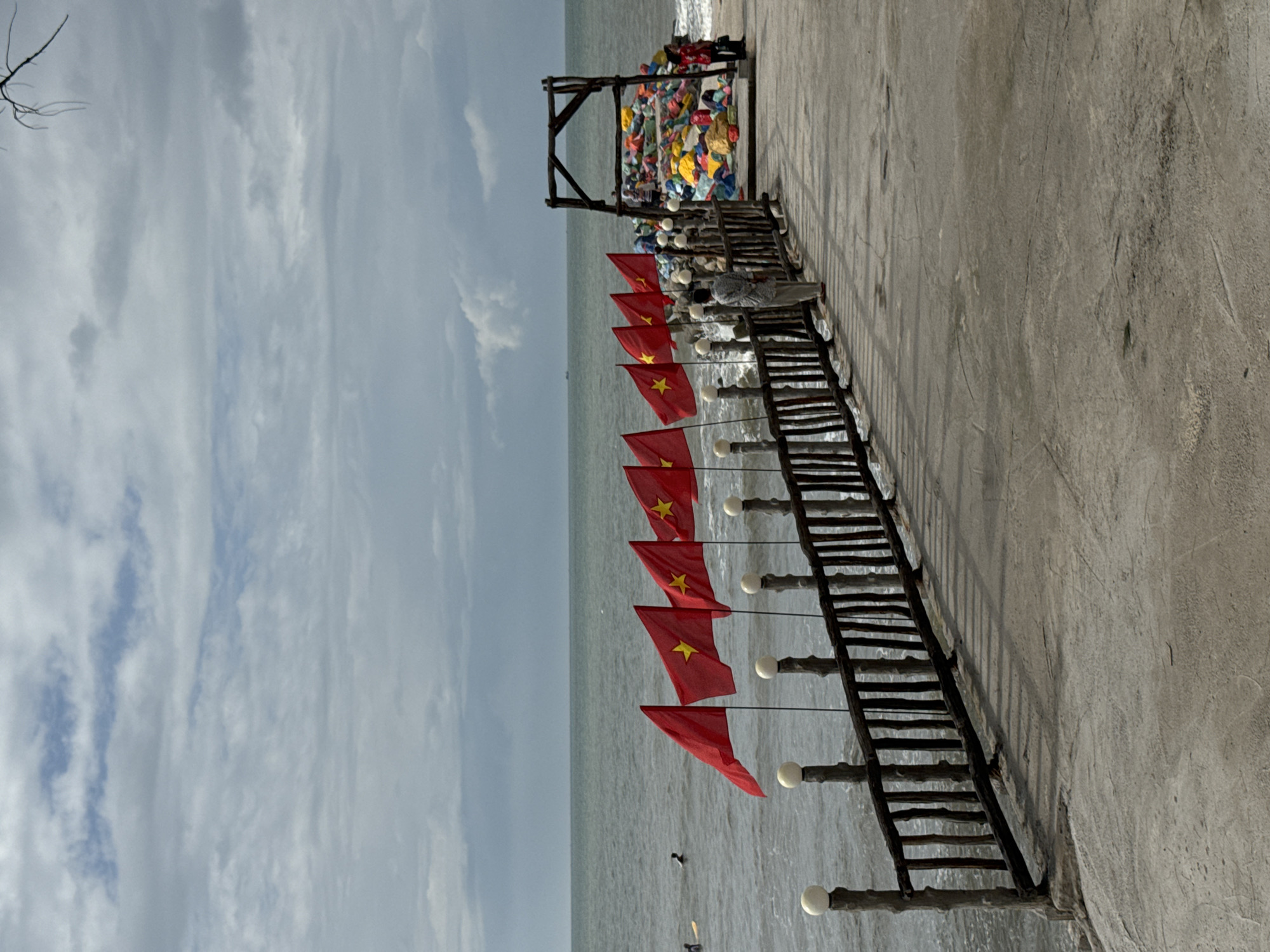 The image depicts a scene on a paved walkway by the sea, likely in Phan Thiết, Vietnam, given the presence of Vietnamese flags. In the foreground, a wooden railing with vertical slats and spherical finials lines the walkway. Beyond the railing, the ocean is visible with a slightly choppy surface and a cloudy sky overhead. Several red flags with yellow stars, specifically Vietnamese flags, are prominently displayed along the railing, fluttering in the breeze.
Further down the walkway, a person is standing and looking out towards the sea. To the right, a wooden arch structure frames a large pile of colorful, stacked bags or cushions. Two individuals are interacting near this pile, one of whom appears to be pointing. In the distance, two surfers are in the water. The overall atmosphere suggests a coastal resort or public area designed for recreation and viewing the sea.