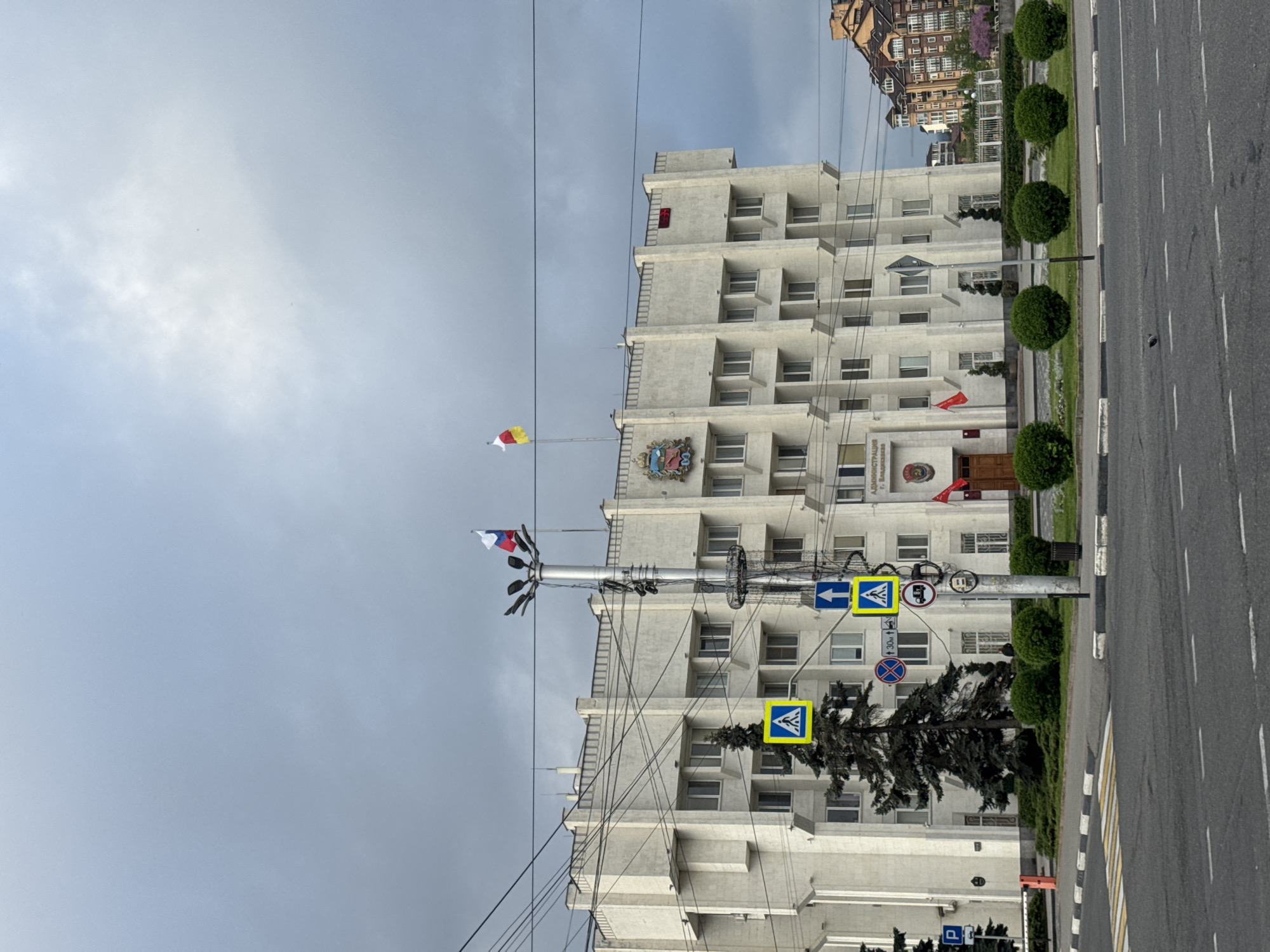 The image shows a building in the city of Akhieli, Georgia. The building is white and has many windows. There are flags flying on the building's roof, including the flag of Georgia and another flag with red, yellow, and white stripes. The building is located next to a street with trees and bushes along the sidewalk. There are several traffic signs visible, including a pedestrian crossing sign. The sky is cloudy.