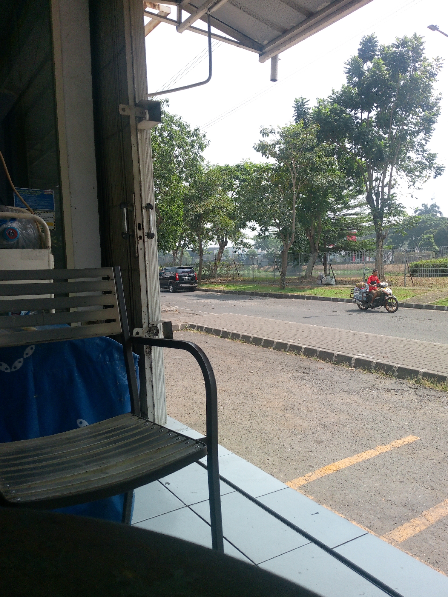 A street scene is depicted, viewed from under an overhang, likely a shop or open-air establishment, in Cikupa, Indonesia. In the foreground, a metal chair with a slatted seat is partially visible, casting a shadow. Behind it, a blue tarp is draped. To the left, a metal rolling door is visible. The street itself is paved with asphalt, bordered by a concrete sidewalk and a metal fence. A black SUV is parked on the side of the road. Further down the street, a person is riding a motorcycle, wearing a red shirt and carrying items secured to the back of the motorcycle. The background features several large trees with green foliage and a bright sky. The overall impression is of a typical day on a moderately busy street.