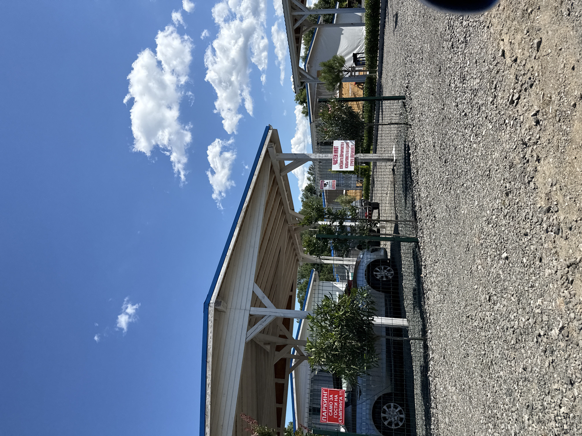 The image depicts an outdoor scene under a bright blue sky with scattered white clouds. In the foreground, a gravel-covered ground leads to a fenced area. Within the fenced area, there is a covered parking structure with a white roof and wooden supports, a silver car parked underneath, and some green foliage. Two red and white signs are visible on posts within the fenced area. The sign on the left reads "ПАРКИНГ САМО ЗА ГОСТИ НА КЪМПИНГА!!!" which translates to "PARKING ONLY FOR CAMPING GUESTS!!!". The sign on the right, partially obscured, contains text including "ЧАСТЕН ИМОТ" ("PRIVATE PROPERTY") and "ЗАБРАНЕНО!!!" ("FORBIDDEN!!!"). In the background, more structures, possibly cabins or other camping facilities, are visible, some with white walls and blue roofs. Trees and more foliage are present throughout the scene. The overall impression is of a campground or a similar recreational facility. The location is identified as being in the city of Tsarevo, Bulgaria.