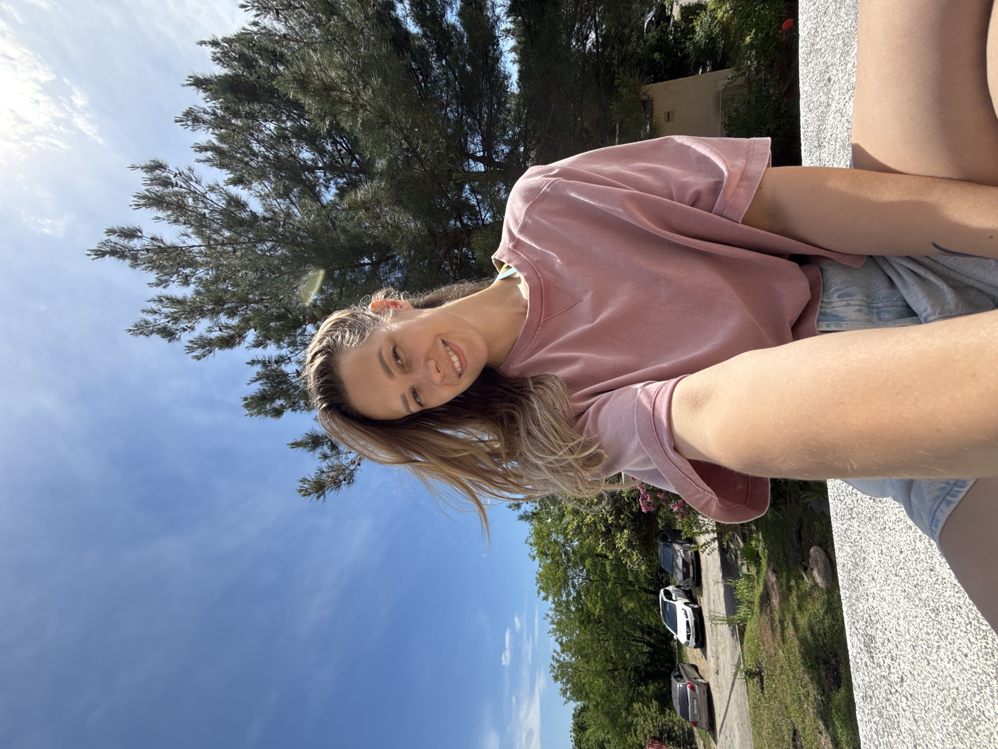 A medium shot shows a young woman with blonde hair and a pink sweatshirt taking a selfie outdoors. She is smiling at the camera. In the background, there are trees, cars, and a bright blue sky with scattered clouds. The location appears to be a parking area with greenery and a building in the distance. The overall scene suggests a warm, sunny day.