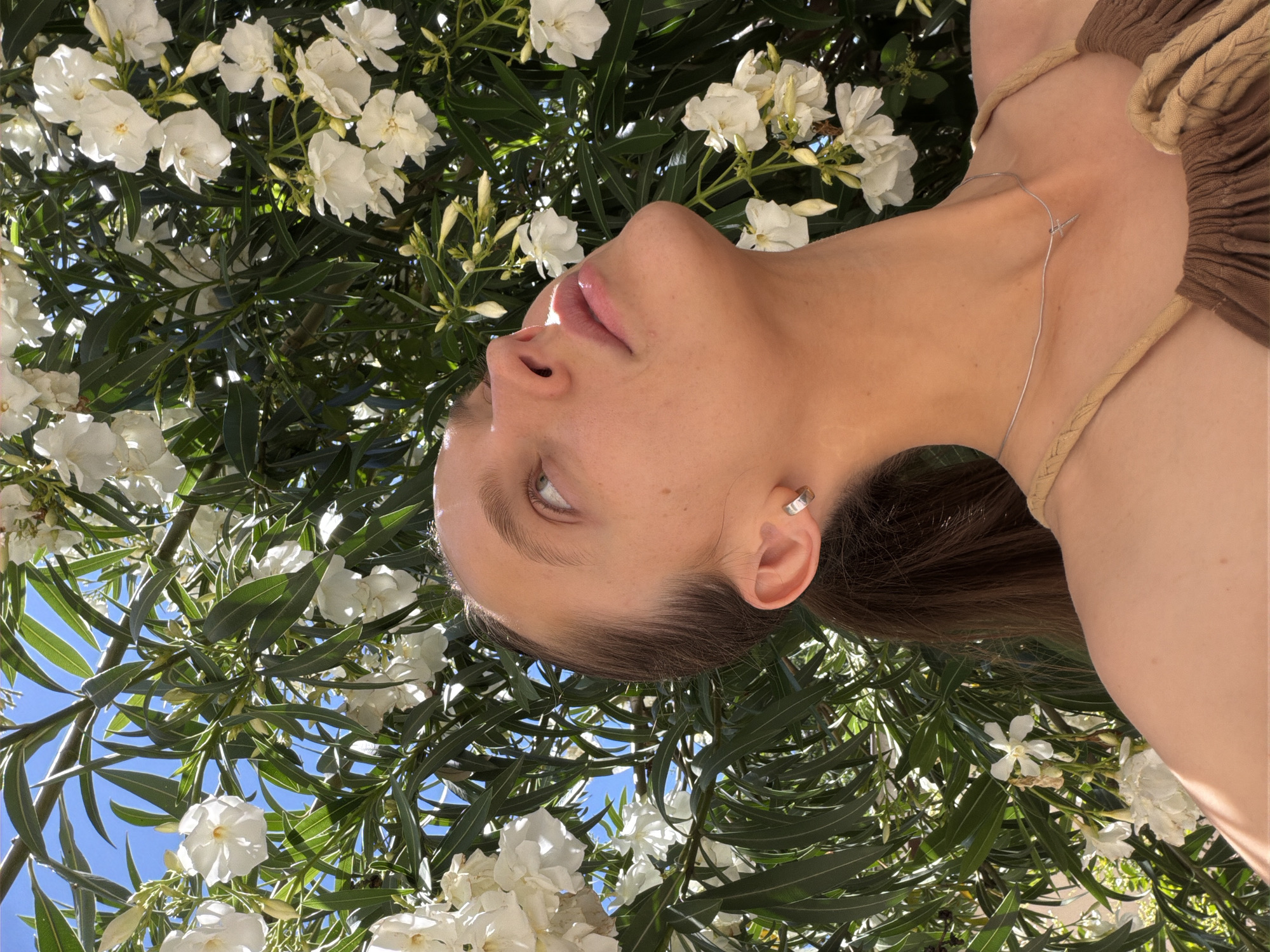 The image, rotated 90 degrees clockwise, presents a close-up portrait of a person positioned against a vibrant natural background. The subject, a young woman, is seen from the chest upwards, with her head tilted back, looking towards the top of the original (unrotated) frame. She has smooth skin, light-colored eyes, and full, slightly glossy lips. A small silver ear cuff is visible on her right ear, and a delicate silver necklace with a minimalist pendant rests on her neck. She is wearing a light brown and tan braided top, and her dark brown hair is pulled back.
The background is dominated by lush green foliage and clusters of white flowers, strongly resembling oleander, indicative of a warm, Mediterranean climate like Budva, Montenegro. Patches of bright blue sky are visible through the leaves, confirming a clear, sunny day. Strong sunlight creates highlights on the person's skin, lips, and the petals and leaves, contributing to a serene and natural aesthetic. There is no visible text in the image.