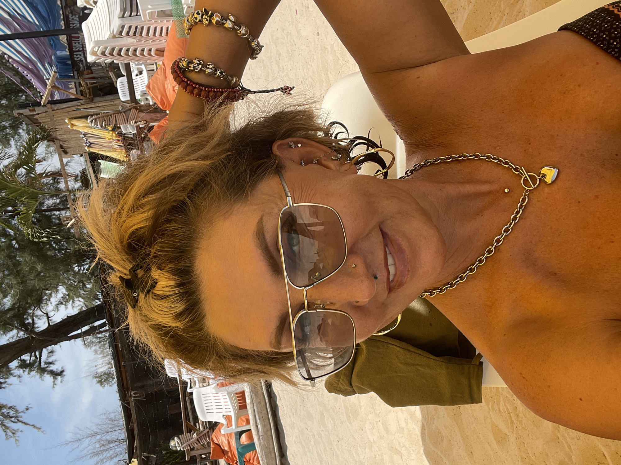 The image shows a woman wearing sunglasses and a gold chain necklace with a heart-shaped pendant, relaxing on a beach in Choeng Thale, Thailand. She is likely at a beach resort, as stacks of white lounge chairs and beach umbrellas are visible in the background. The location appears to be a sunny, tropical beach.
