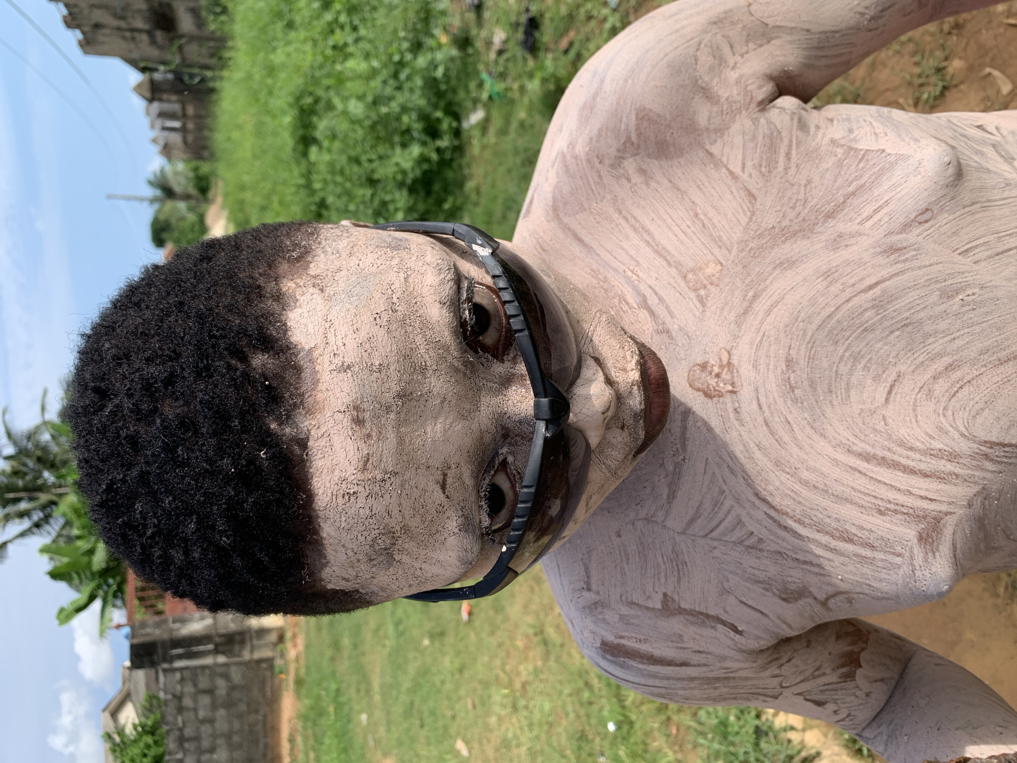 A close-up shot shows the face and upper body of a person covered in a light-colored paste, possibly clay or paint. The person has dark, curly hair and is wearing dark-lensed goggles. The paste is applied in textured strokes, with some areas appearing smoother than others. The person's eyes are visible through the goggles, looking directly at the camera. The background is blurred, showing green foliage, a dirt path, and hints of buildings in the distance. The lighting suggests it is daytime. The scene appears to be outdoors, in a rural or semi-rural setting in Ikorodu, Nigeria. The overall impression is one of a ritual or artistic performance.