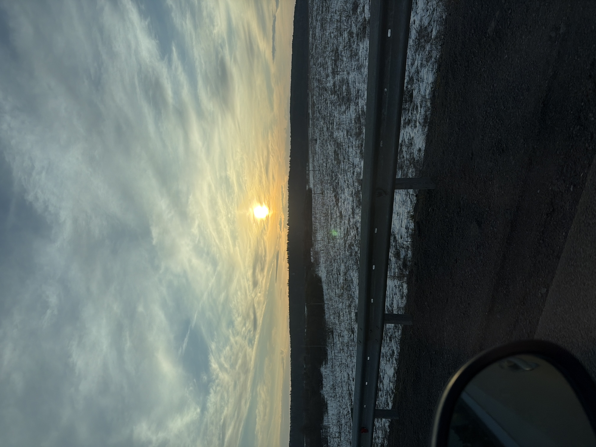 The image shows a view from a vehicle traveling on a road near Chernovskoye, Russia. The sky is predominantly blue with wispy white clouds tinged with yellow from the setting sun, which is low on the horizon. A dark treeline separates the sky from a snow-covered field, visible on the right side of the frame. A metal guardrail runs along the road, and the dark asphalt surface is prominent. A portion of the vehicle's side mirror is visible in the lower right corner. There are no people visible in the frame.