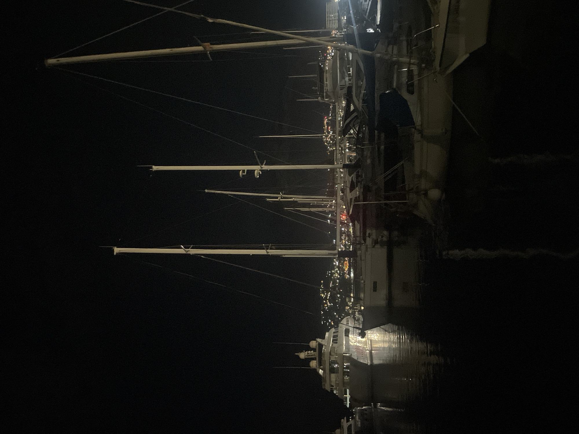 The image depicts a nighttime scene at a marina in Bar, Montenegro, with several yachts and sailboats docked. The foreground is dominated by the dark water of the marina, reflecting the lights of the boats and the distant city. Several tall masts of sailboats are visible, silhouetted against the night sky. In the mid-ground, a large, illuminated luxury yacht is prominent, showcasing its multiple decks and windows. Behind the boats, the hills of Bar are visible, dotted with numerous lights from the city and buildings, creating a twinkling effect. The overall impression is of a serene and affluent harbor at night.