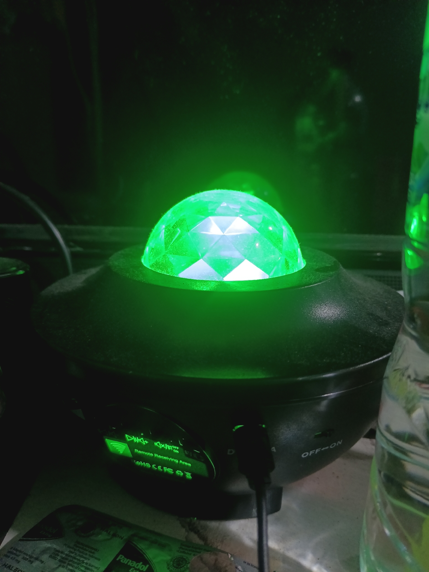 An illuminated green disco ball is perched atop a black, UFO-shaped device. The device has a glowing green control panel with symbols and text, including "Remote Receiving Area" and "OFF<->ON". A black USB cable is plugged into the side of the device. To the right, a partially visible clear plastic bottle with a green liquid is present. The background is dark and appears to be an indoor setting, possibly a room in Sukamekar, Indonesia. There are no people or animals in the frame.