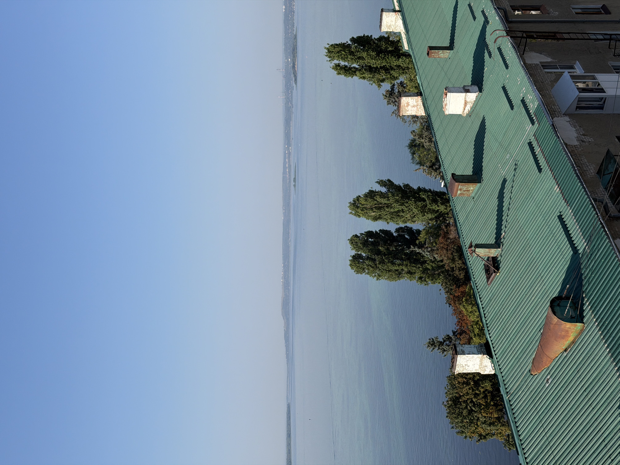 The image captures a view from a rooftop in Saratov, Russia, overlooking a wide river under a clear blue sky. The foreground is dominated by a green, corrugated metal roof adorned with several chimneys, some showing signs of age and rust. A prominent rusty, conical object lies on the roof, its purpose unclear. To the left, lush green trees with hints of autumnal colors suggest a transition in season. Further in the distance, the expansive, tranquil river stretches towards a hazy horizon, where a thin strip of land with buildings and industrial structures can be discerned. The overall scene conveys a sense of quiet observation, looking out over the city and its natural surroundings.