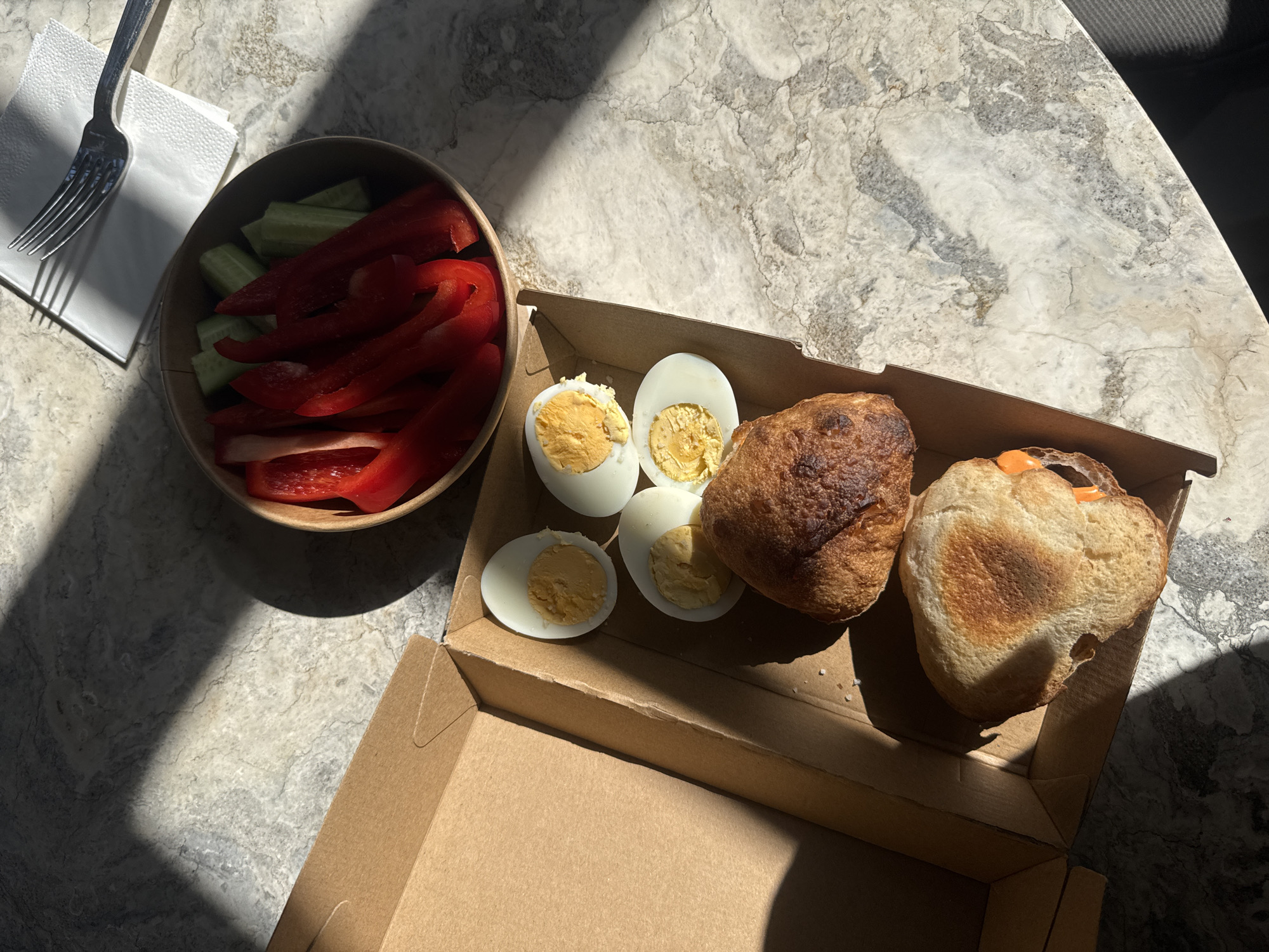 A top-down shot shows a prepared meal likely purchased for takeout. The meal consists of a round cardboard bowl filled with red bell pepper strips and green cucumber slices. Next to it is a small, open cardboard box containing two halved boiled eggs and two pieces of bread. A fork and a folded napkin are visible in the upper left corner of the frame. The meal is positioned on a marble-patterned surface, likely a table. The lighting suggests it is daytime, with shadows cast across the scene. Given the context, the location is estimated to be in Tel Aviv, Israel, though no definitive markers verify the exact placement. No people or interactions are directly visible. The focus is strictly on the prepared meal and its immediate surroundings.