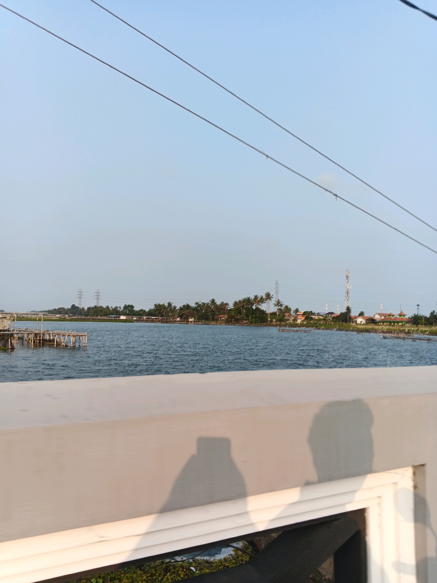 A wide shot captures a serene waterscape with a bright, clear sky. In the foreground, a white railing runs horizontally, casting a shadow of a person holding a phone, suggesting a selfie is being taken. Beyond the railing, a calm body of water stretches towards a tree-lined horizon. A wooden pier or structure extends into the water on the left side of the frame. The opposite shore is dotted with houses, palm trees, and several tall communication towers. The scene appears to be a picturesque view of the Cilacap coast in Indonesia, known for its beaches and waterfront attractions. The water is a deep blue, and the sky is a pale blue, indicating a clear day.