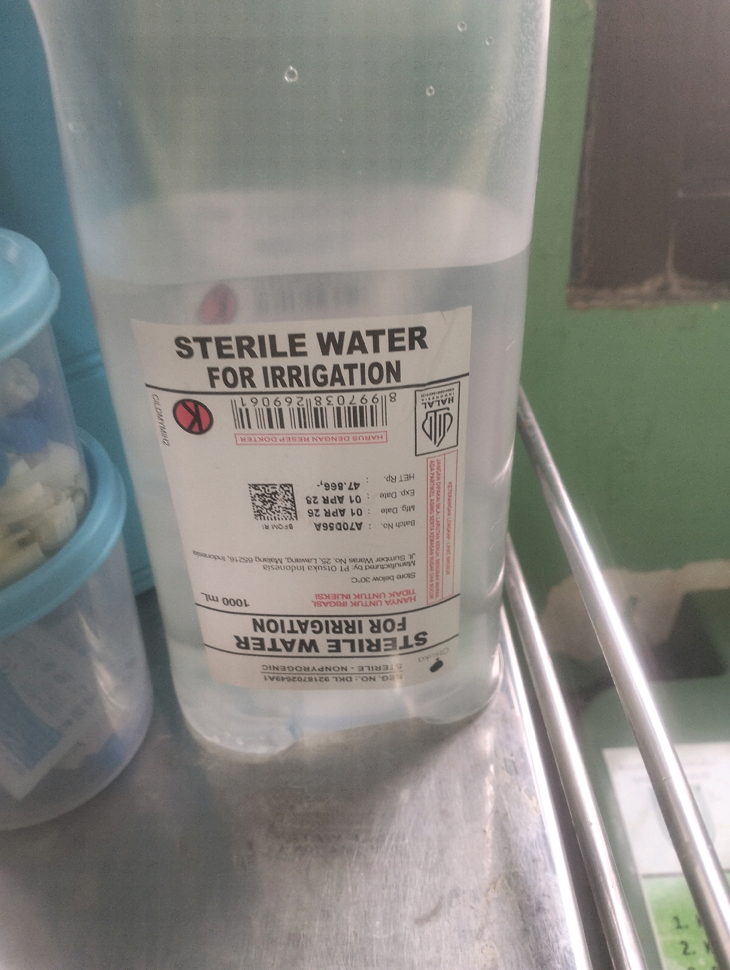 The image shows a close-up of a plastic bottle of "STERILE WATER FOR IRRIGATION". The bottle is mostly transparent, filled with clear liquid. A white label with black text is adhered to the front of the bottle. The label contains product information, including batch number, manufacturing date, expiration date, and volume (1000 mL). The text also indicates that the product is manufactured by Otsuka Indonesia. The bottle appears to be sitting on a metallic surface, possibly a tray or table. There are other medical-related items visible in the background, including a blue plastic container and some chrome-colored railings. The environment appears to be a medical setting. There is no discernible indication of the specific location in Air Jamban, Indonesia, beyond the manufacturing origin of the product.