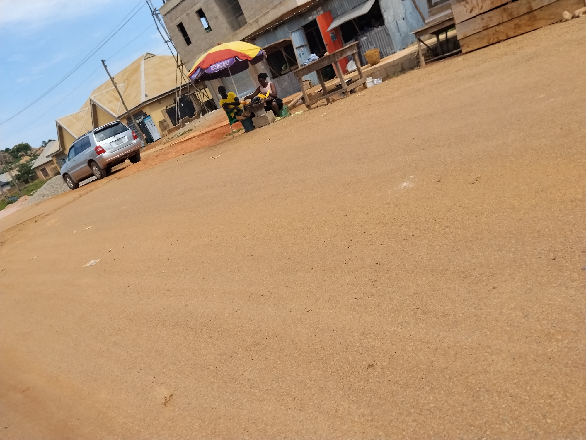 In Nabatum, Nigeria, a dusty, unpaved road cuts through a semi-urban landscape under a clear sky. On the left, a silver SUV is parked on the side of the road. To the right, two individuals are seated under a colorful umbrella near a makeshift stall. One person, wearing a yellow shirt, appears to be engaged in cooking over a charcoal grill. The other person, in a pink top, is seated next to them. Behind the stall, a wooden table and stacked planks of wood are visible, along with some corrugated metal structures and a basket. The buildings in the background appear to be a mix of under-construction and completed structures. The overall scene depicts a typical street market or roadside vendor setting.