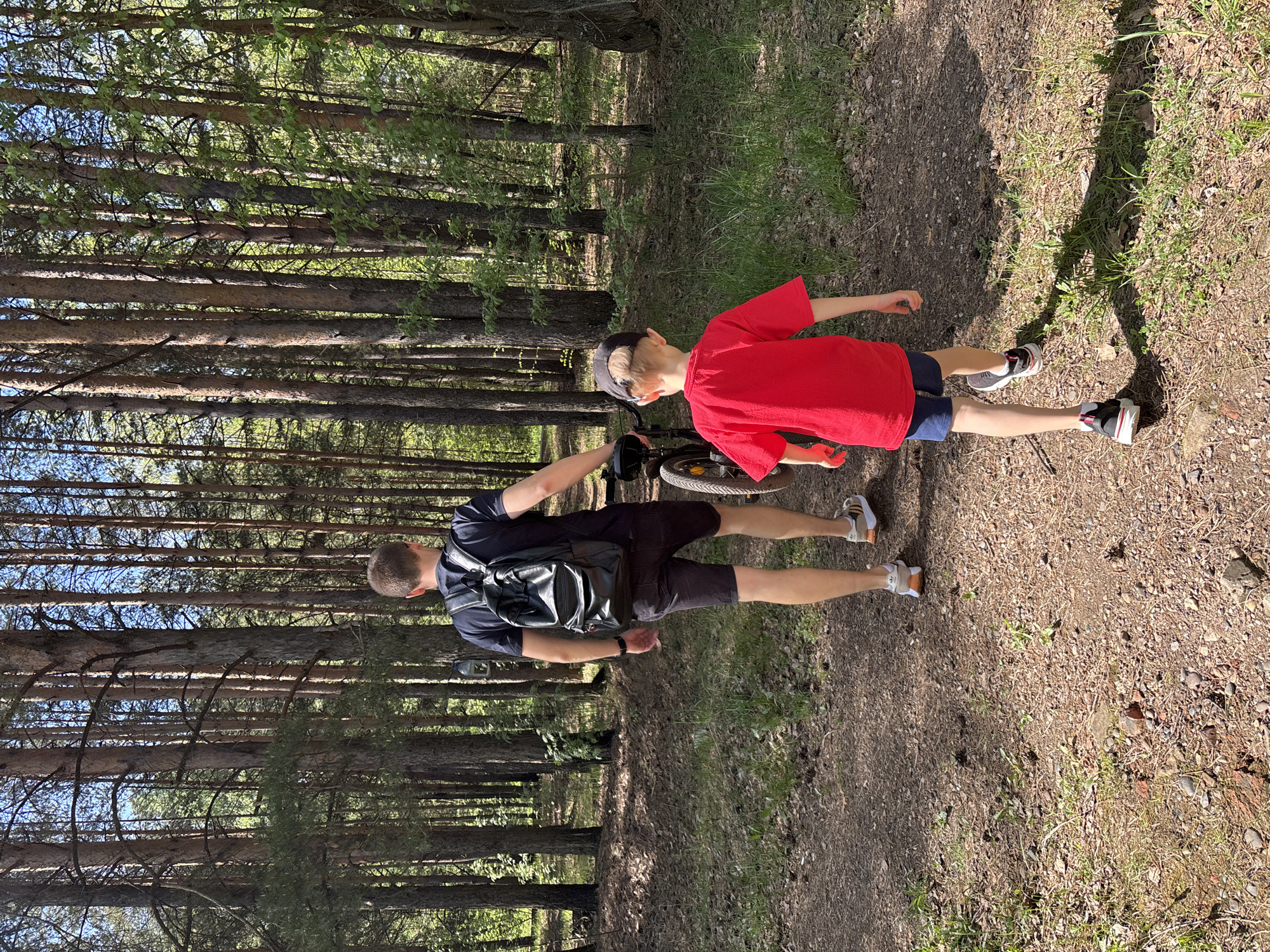 The image shows a forest scene, likely a park or wooded area. A man and a boy are walking on a dirt path through the trees. The man is wearing a dark shirt and shorts, with a backpack. The boy is wearing a red shirt and dark shorts, and appears to be holding a bicycle. The trees are tall and dense, creating a partially shaded environment. The ground is covered in dirt and some grass. Based on the location provided, Chaikovsky, Russia, the scene is likely within a forested or park area in the city.