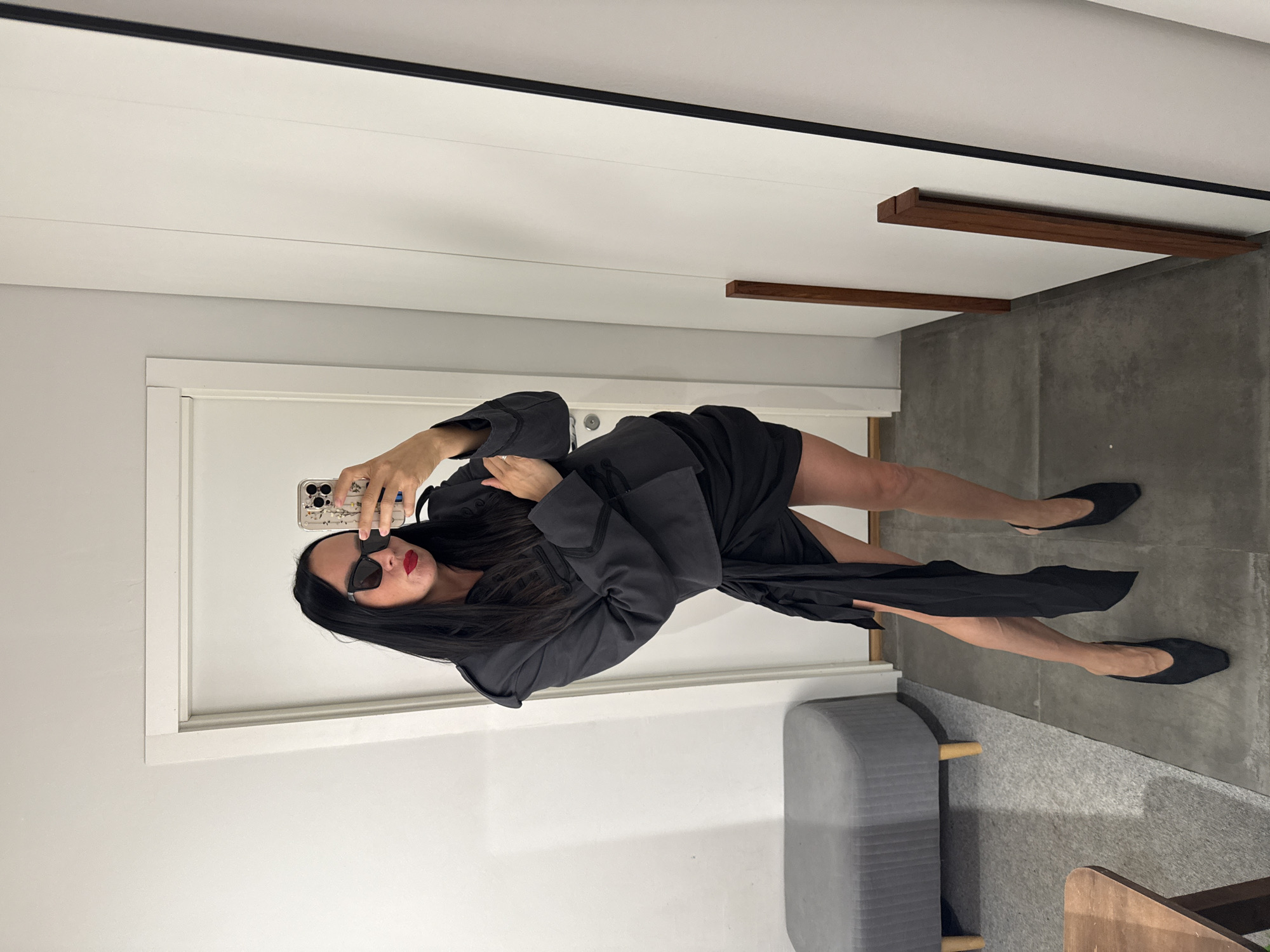 A woman stands in a room, taking a mirror selfie. She has long, dark hair and is wearing large, black, rectangular sunglasses, a dark grey jacket with epaulets and ornate trim, a black miniskirt with a draped fabric detail, and black pointed-toe slingback heels. Her lipstick is bright red. She is holding a smartphone, which is encased in a decorative case featuring floral patterns. The room appears to be a dressing room or a fitting room, with a large mirror on the right wall, a white door on the left, and a grey upholstered bench with wooden legs to the left of the door. The floor is a dark grey concrete. The scene is well-lit.