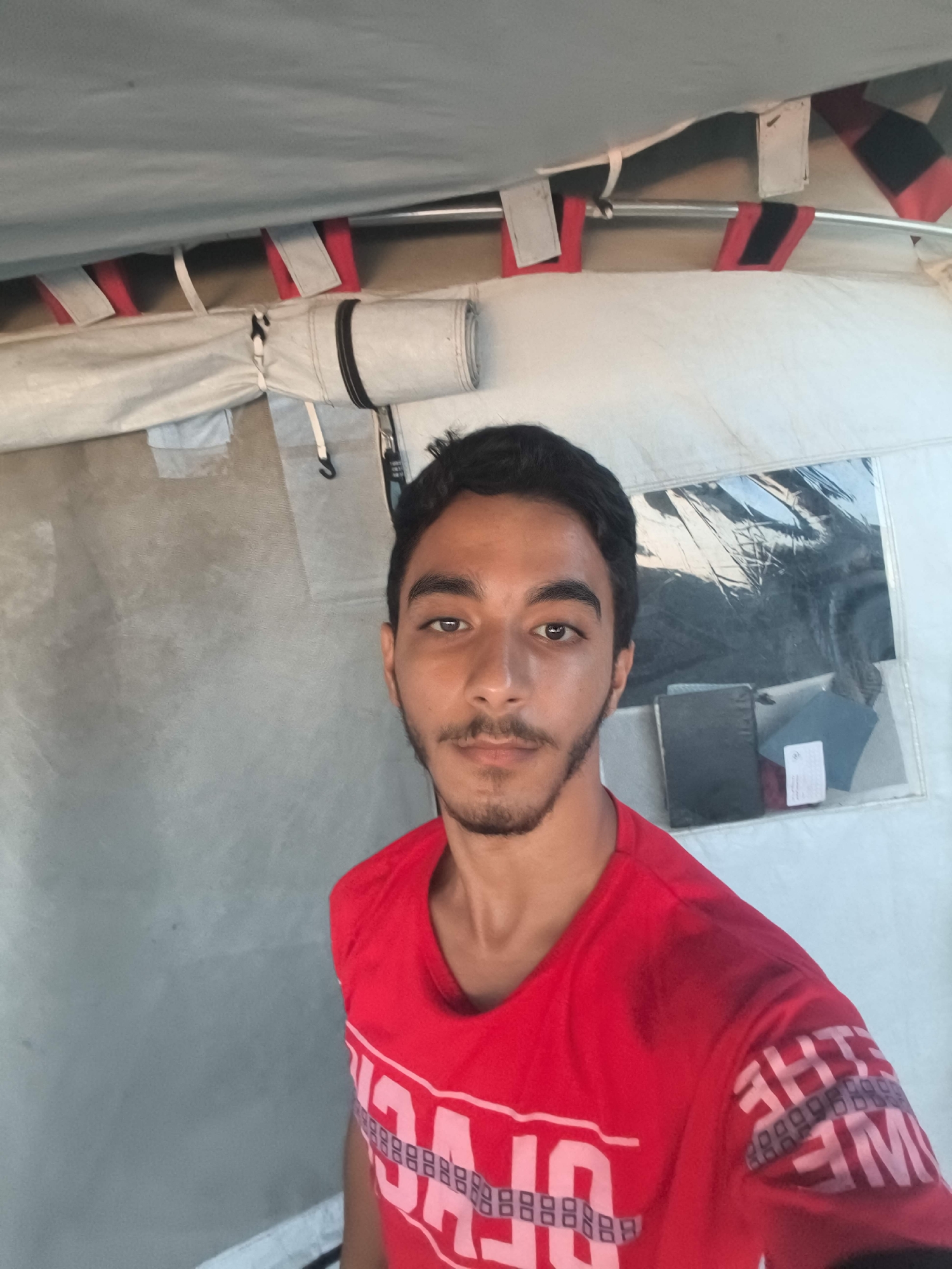 A man with dark hair and a beard is taking a selfie in a tent. He is wearing a red t-shirt with white text. The tent appears to be made of canvas and has a clear plastic window through which books and papers are visible. The background is grey, and there are red and black straps visible on the tent's ceiling.