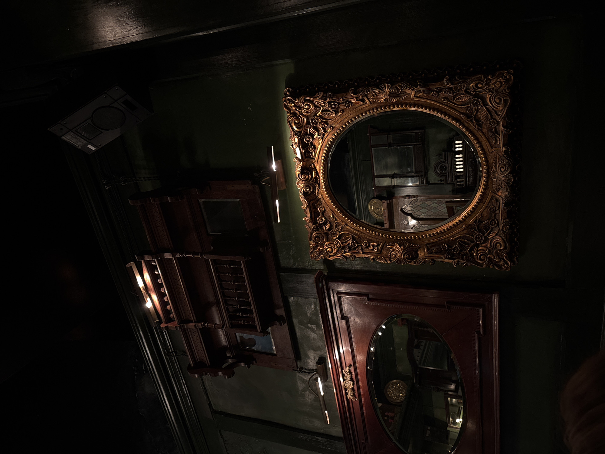 The image depicts a dimly lit interior, possibly a bar or restaurant in Moscow, Russia, given the style of the decor and the presence of ornate mirrors and sconces on dark green walls. An elaborate gold-framed round mirror hangs on the wall, reflecting the interior space and a portion of a counter with objects on it. Below it is a wooden-framed oval mirror reflecting a similar scene. To the left of these mirrors is a tiered wooden structure that appears to be a bar or display unit. Near the top left corner of the frame is an air conditioning unit mounted on the ceiling. The lighting is subdued, with sconces providing the main source of illumination. The overall impression is of an old-world or vintage-style establishment.